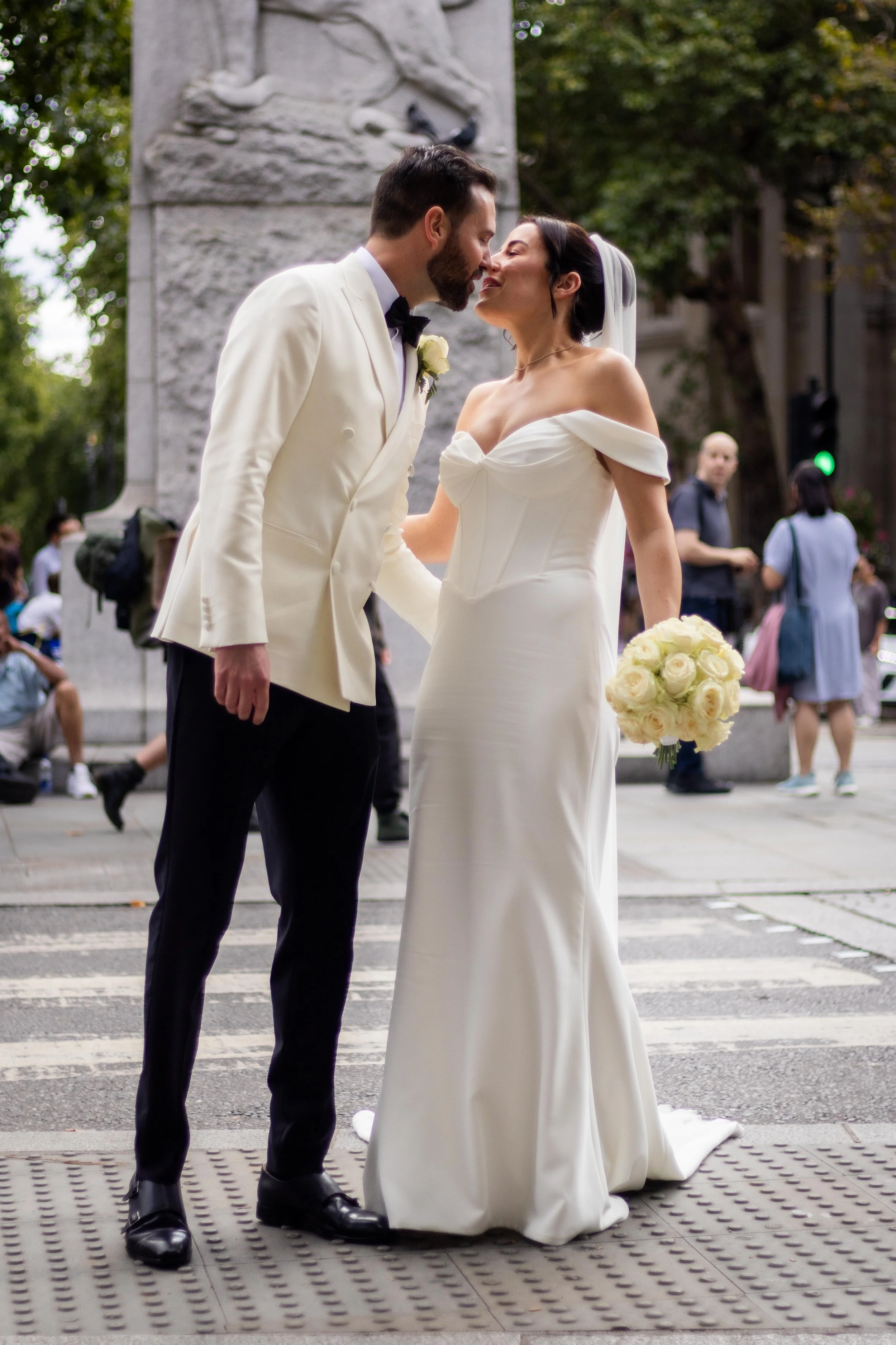 A bride and groom are standing close together on a city street, leaning in for a kiss. The groom wears a white tuxedo jacket, black trousers, and a black bow tie. The bride wears an off-the-shoulder white wedding gown and holds a bouquet of white ros