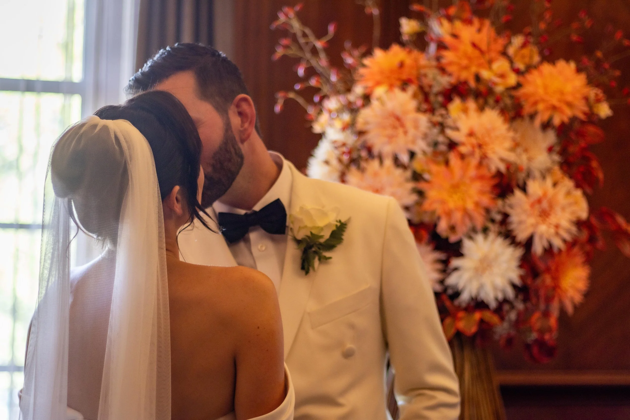A bride and groom sharing an intimate moment during their wedding ceremony, with the bride wearing a veil and the groom dressed in a white tuxedo with a black bow tie, standing in front of a large floral arrangement of orange, white, and pink flowers
