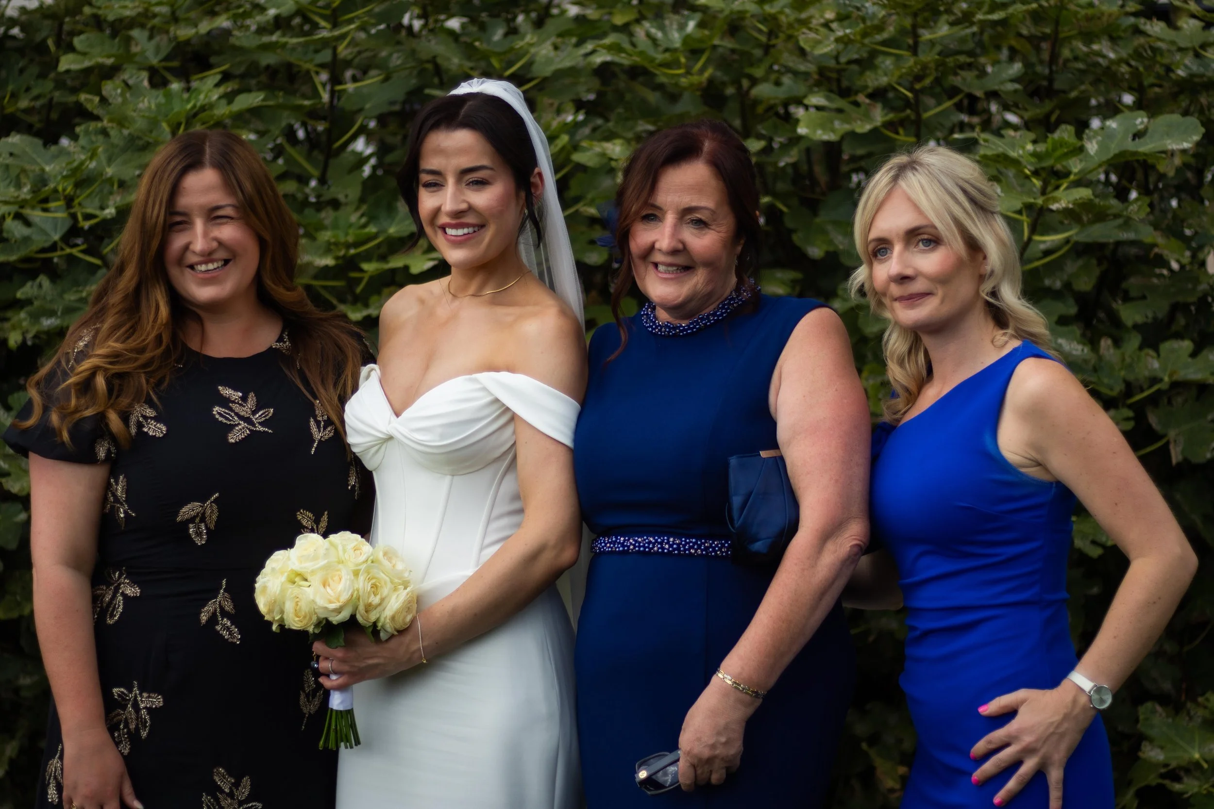 Four women standing outdoors, with green foliage background. One woman in a white wedding dress holding a bouquet of white roses. The other women are dressed in black and blue dresses, smiling and posing for a photo.