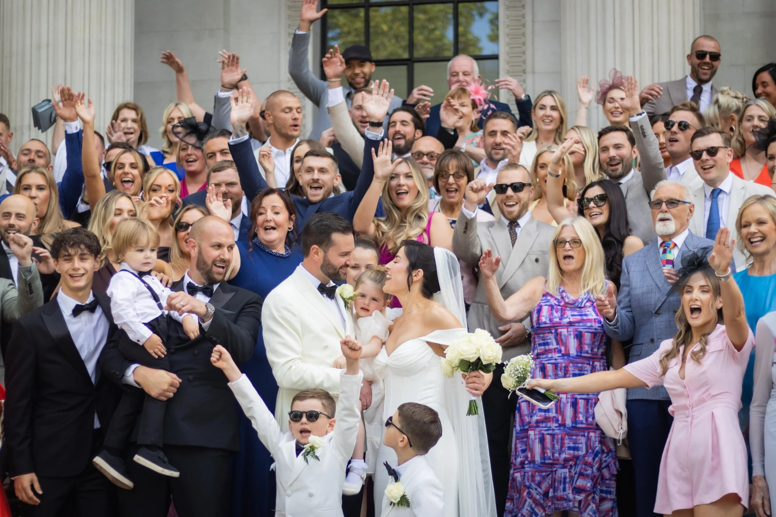 A large wedding celebration with the bride and groom in the center surrounded by family and friends, many raising their hands and smiling outdoors.