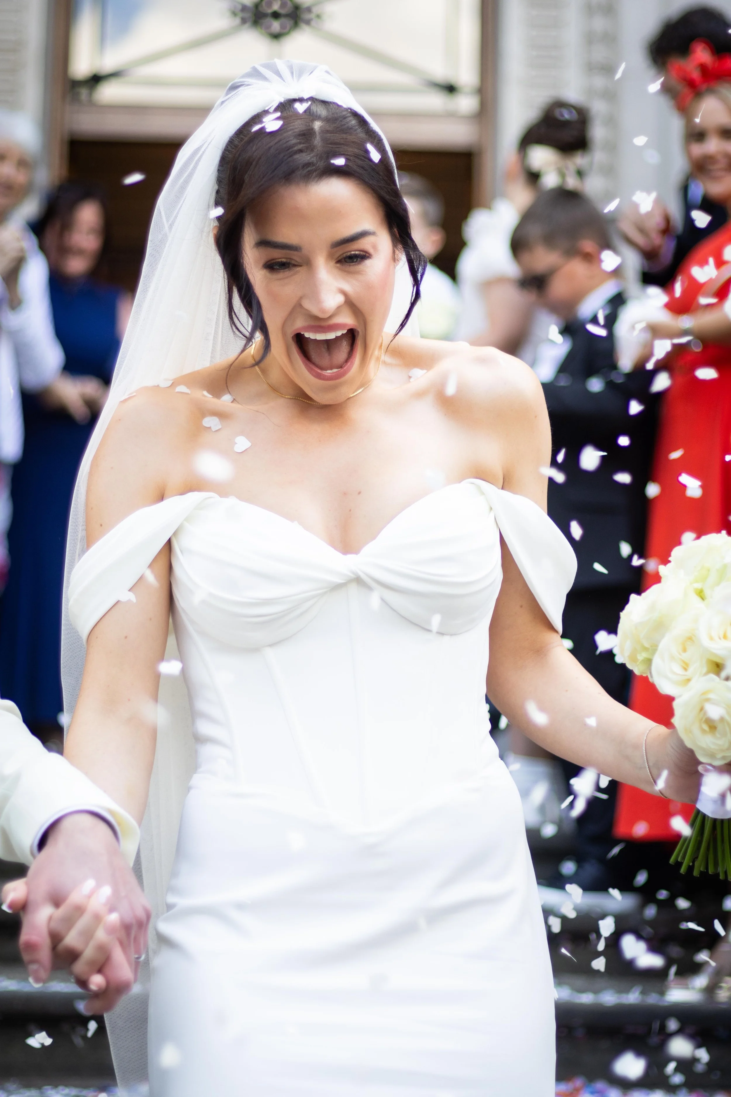 A bride in a white wedding dress and veil celebrating with her wedding bouquet, surrounded by confetti and wedding guests in the background.