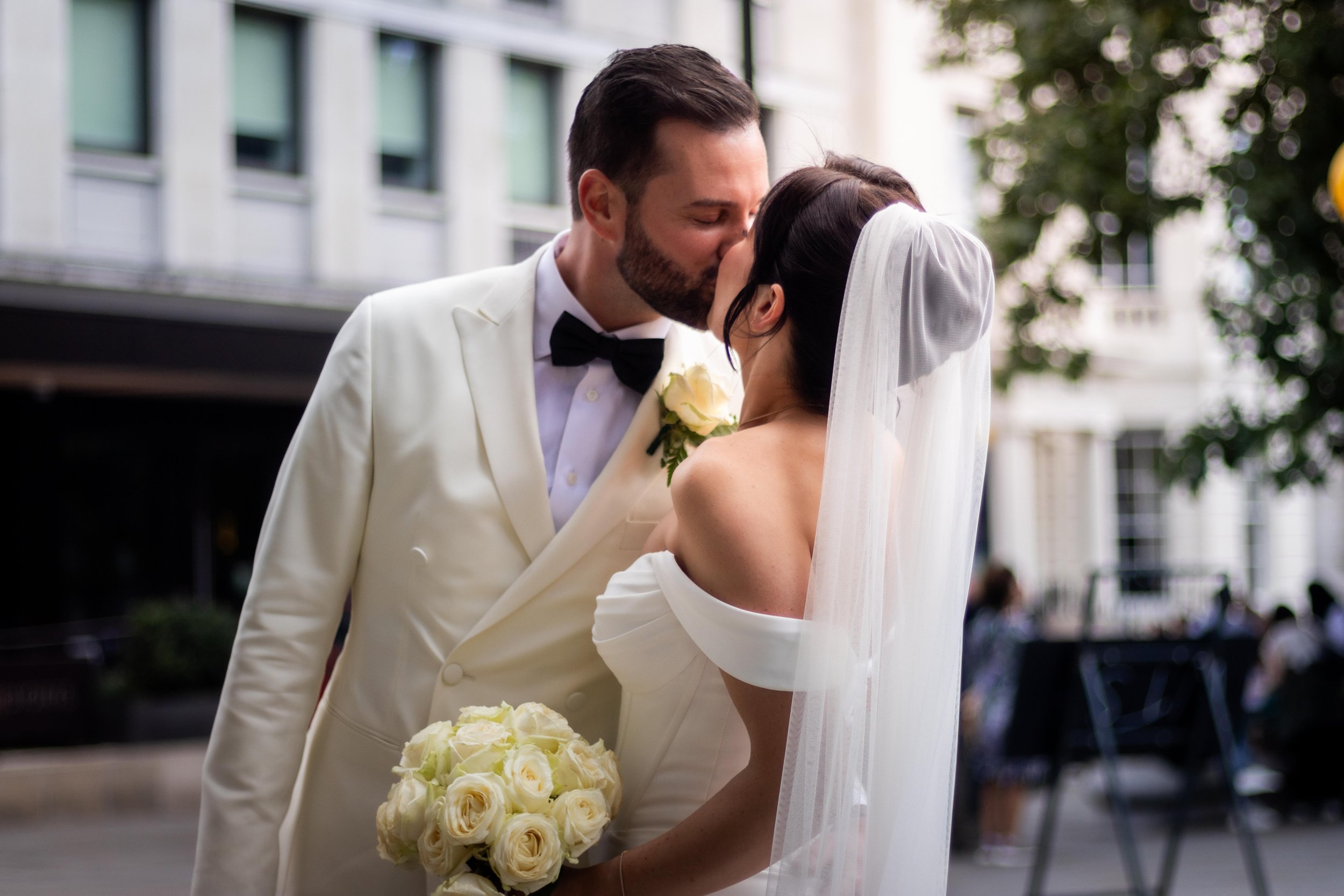 A newly married couple sharing a kiss outdoors in an urban setting, the groom wearing a white tuxedo with a black bow tie and holding a bouquet of white roses.