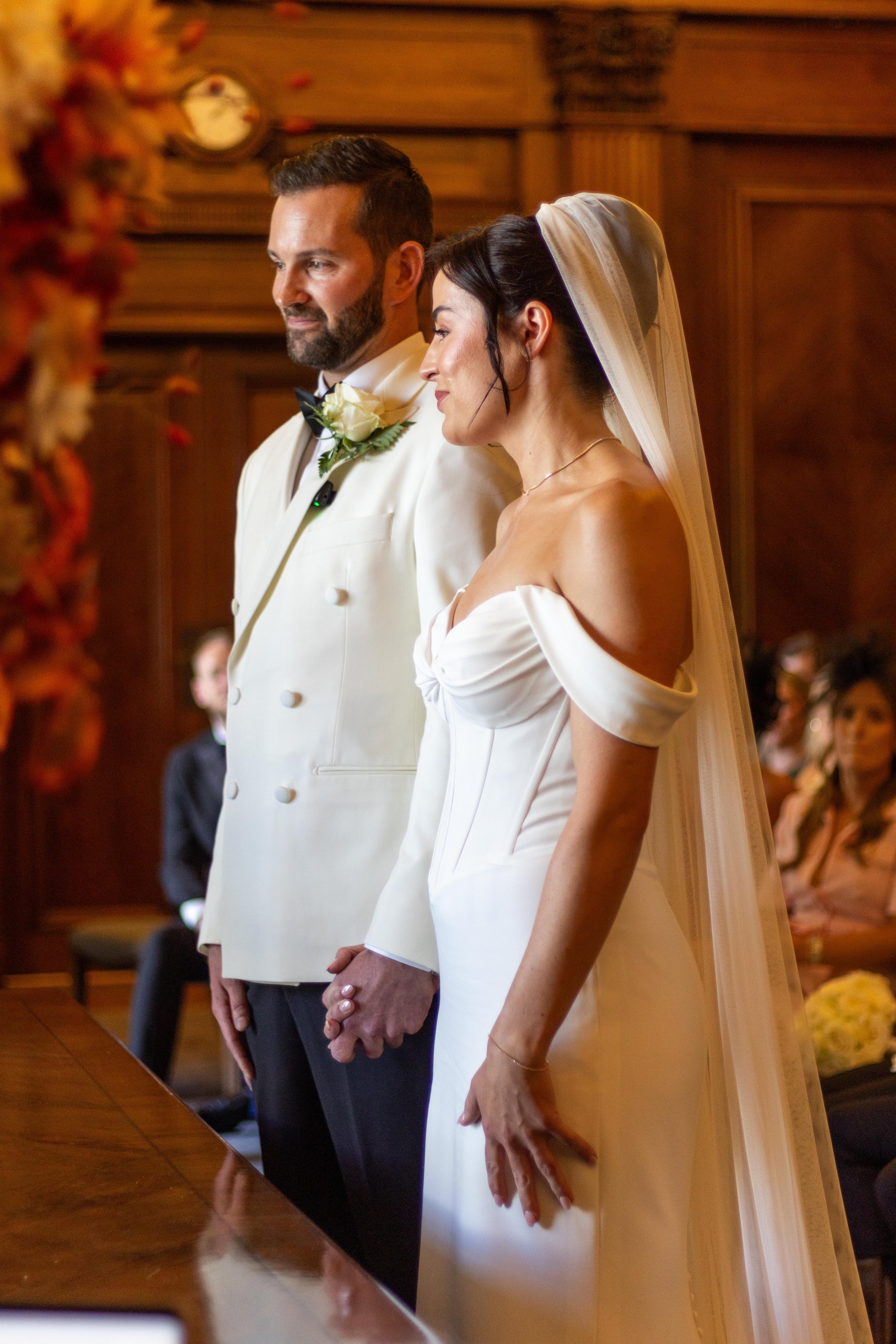 A bride and groom standing hand in hand during their wedding ceremony, with the bride in a white off-shoulder gown and veil, and the groom in a white tuxedo jacket with a black bow tie, in a wood-paneled room with seated guests in the background.