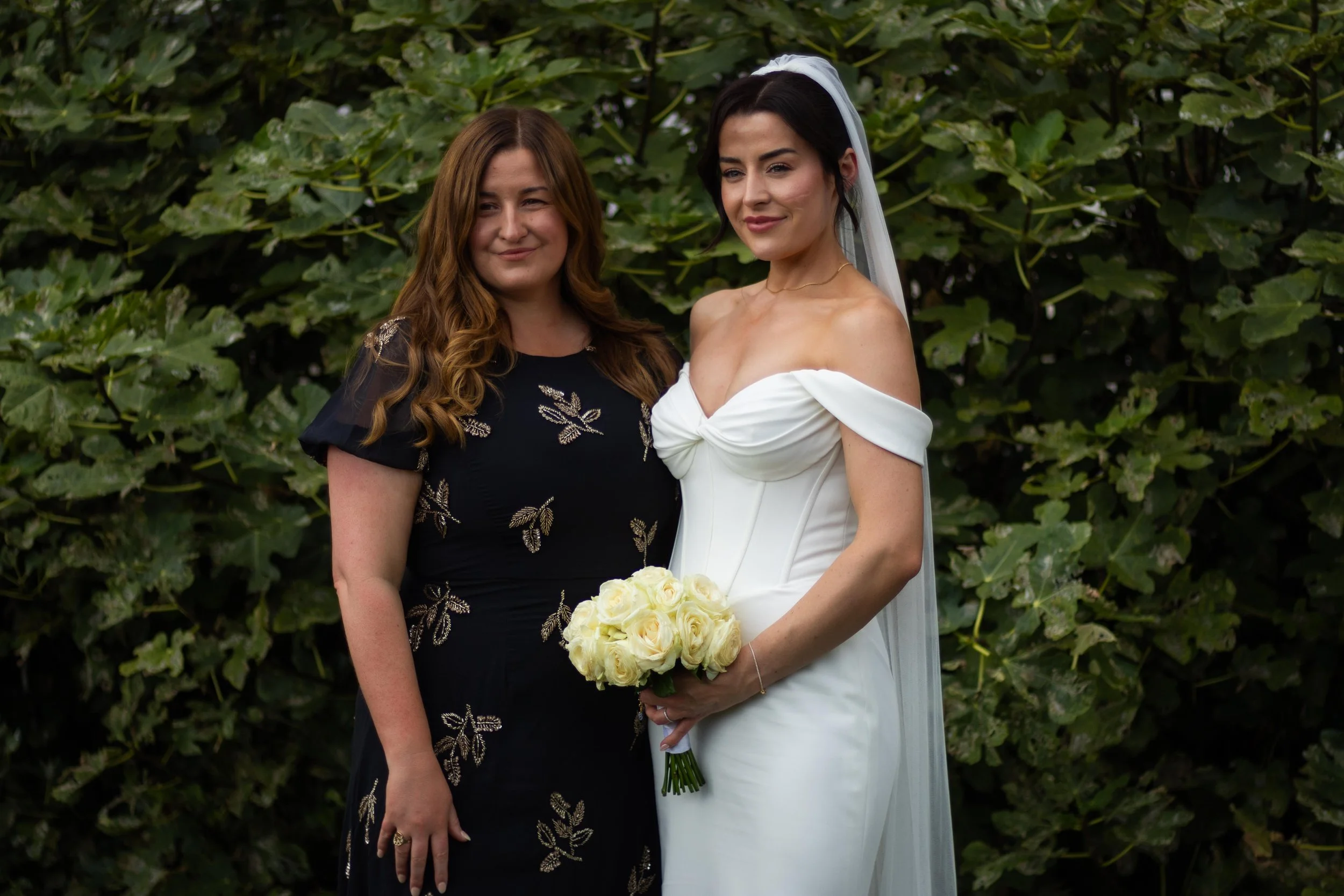 A bride in a white dress holding a bouquet of white roses standing next to a woman in a black dress with gold embroidery, posing outdoors in front of green foliage.