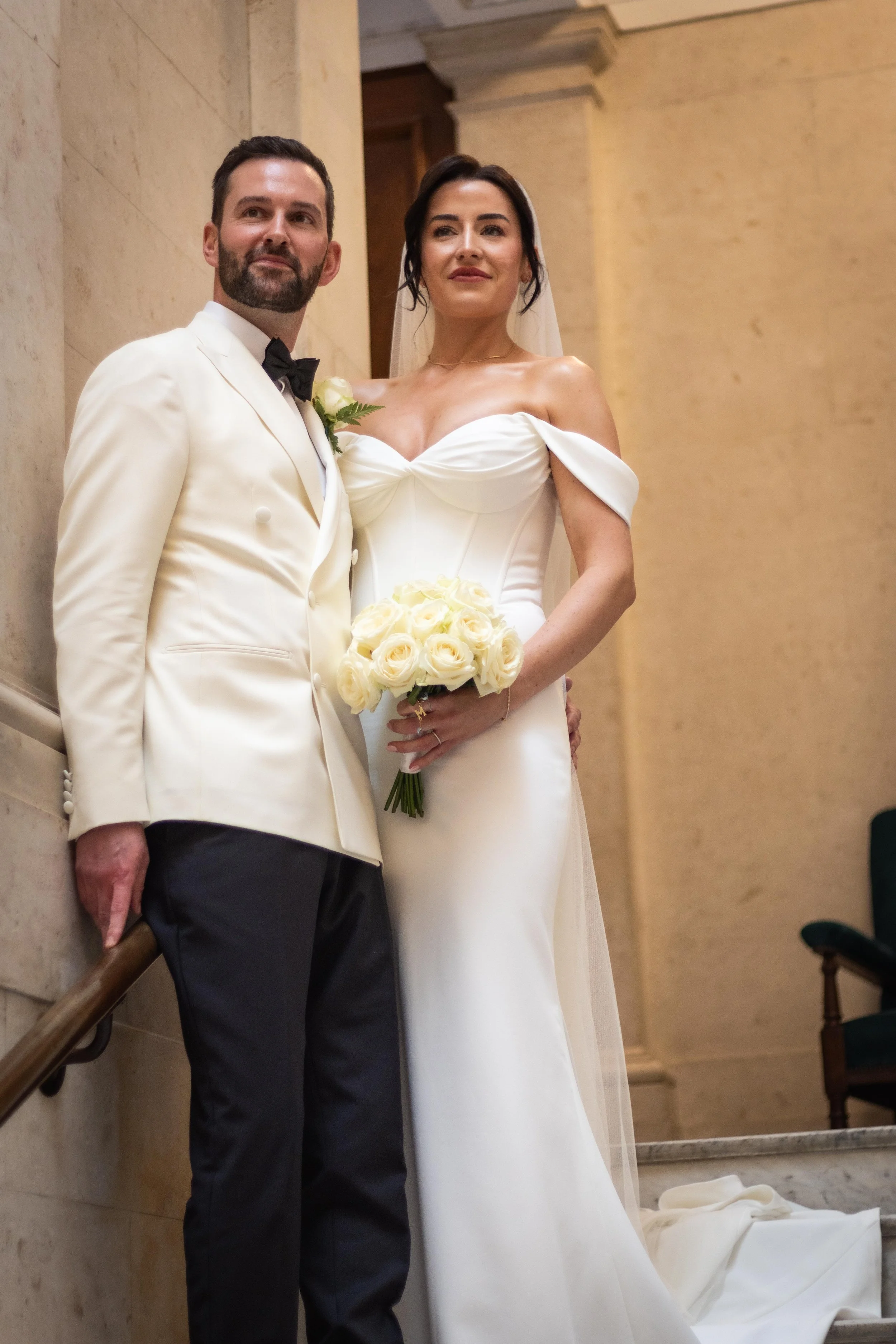 A bride and groom standing together indoors during their wedding, with the bride holding a bouquet of white roses.