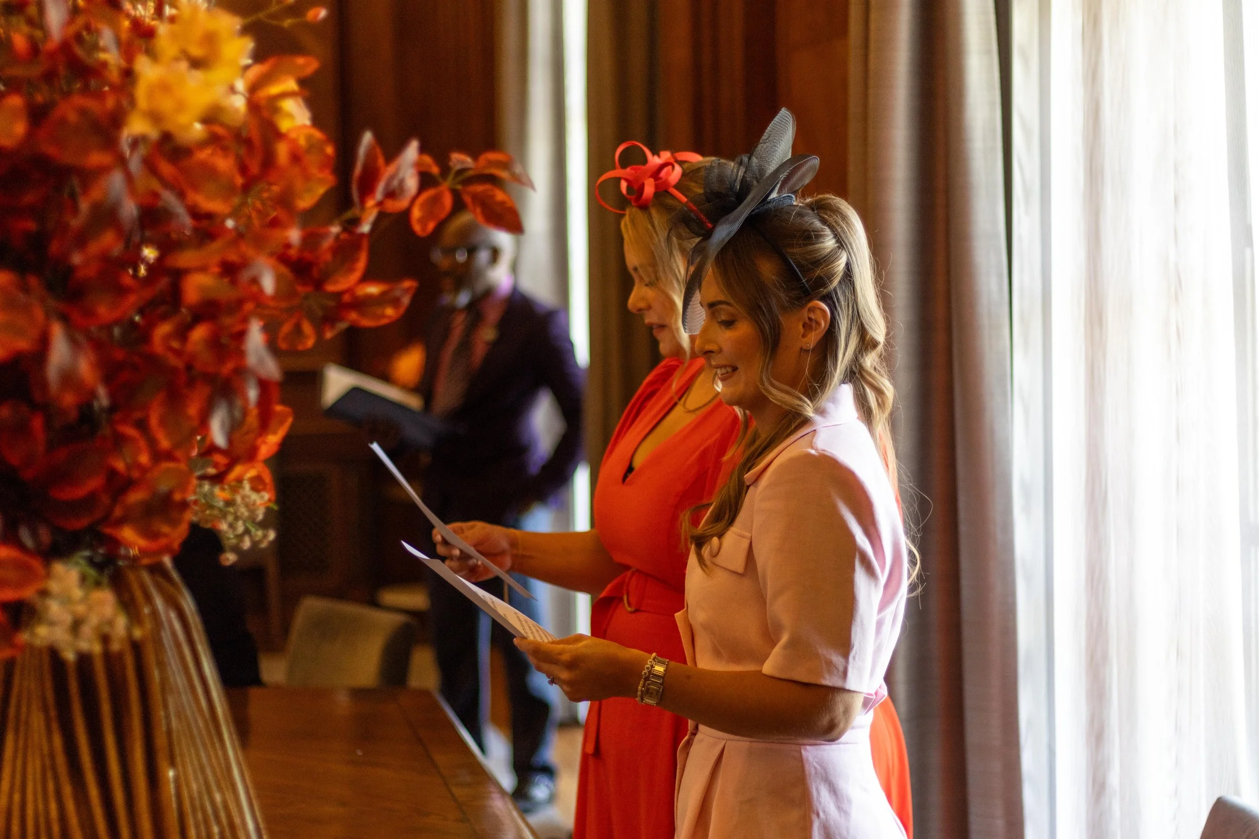 Two women in pink and orange dresses looking at papers, wearing fancy hats, in a warmly lit room with curtains and a large floral arrangement.