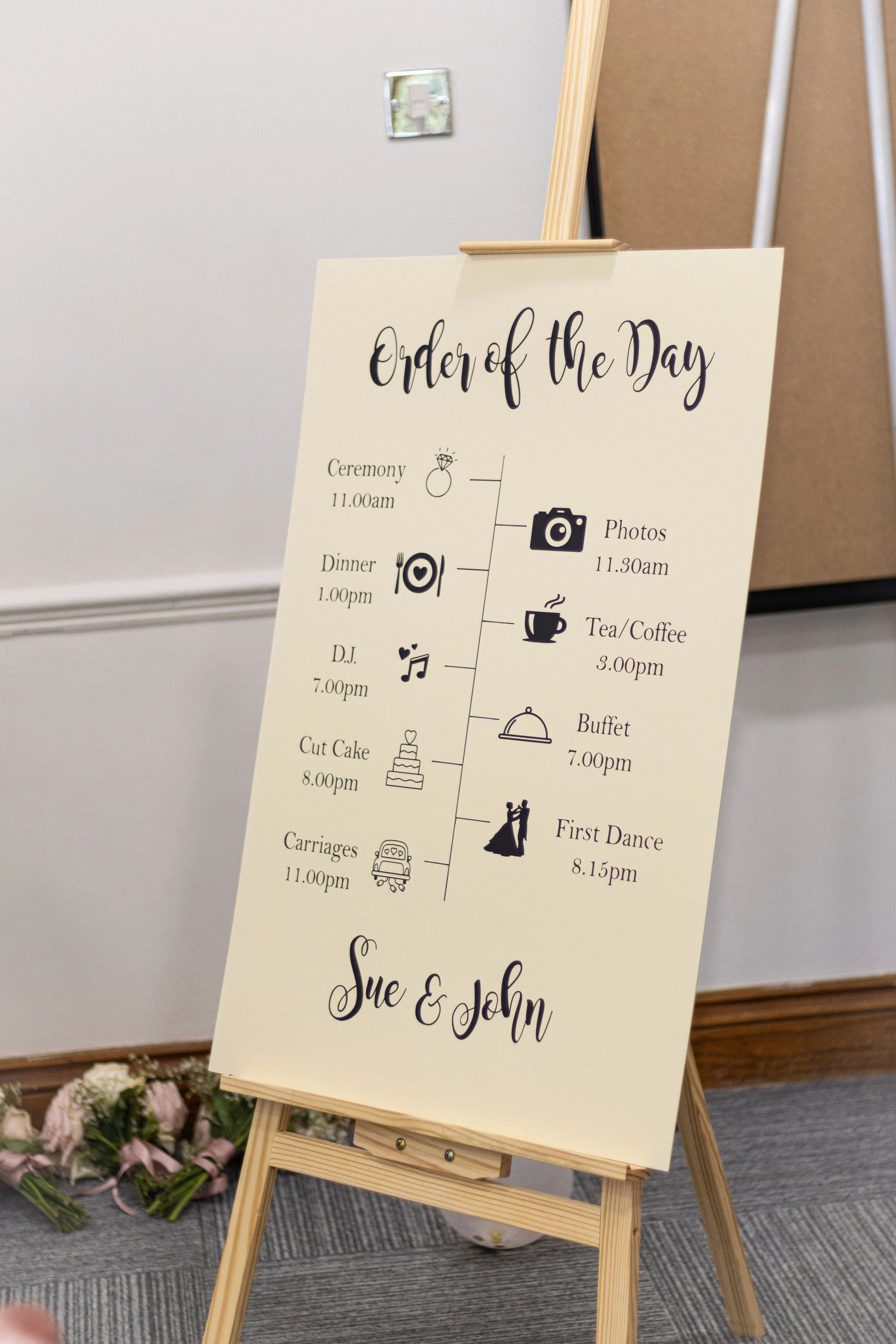 Wedding program sign with schedule of events for Sue and John, including ceremony, photos, dinner, tea/coffee, buffet, first dance, carriages, DJ, and cut cake, with icons and times.
