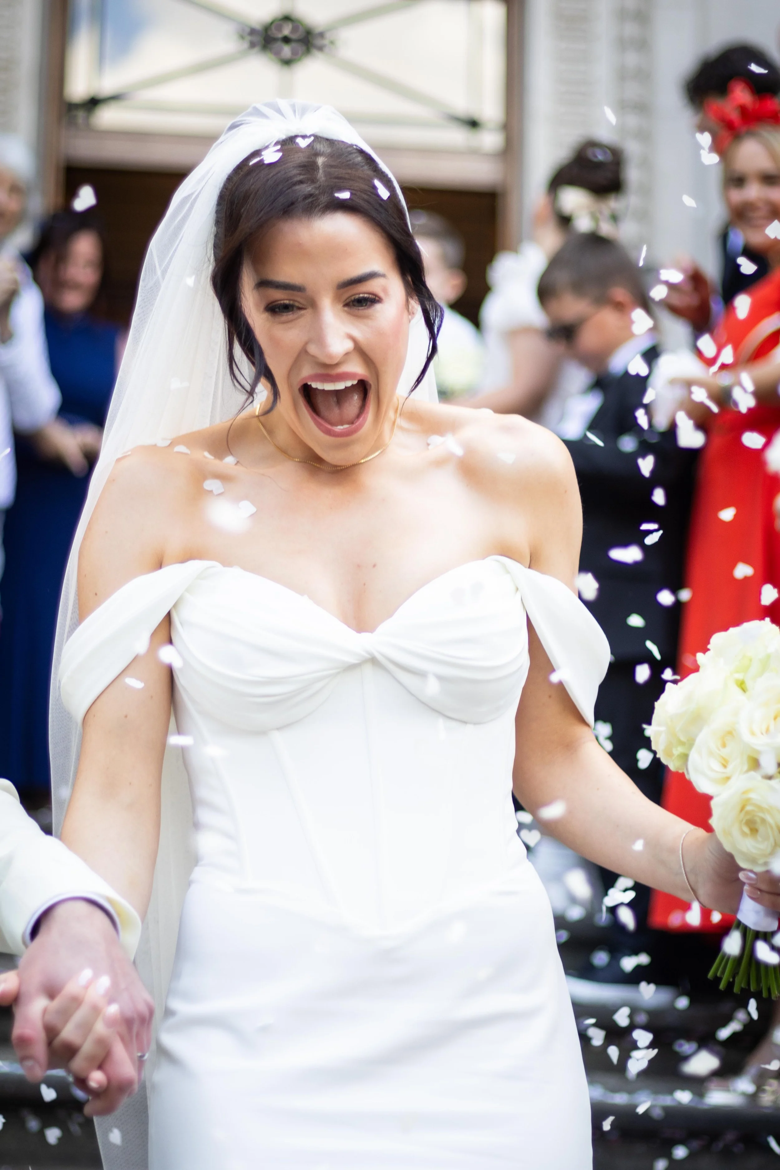 A bride in a white wedding gown with a veil and bouquet, expressing joy as confetti is thrown during her wedding celebration.