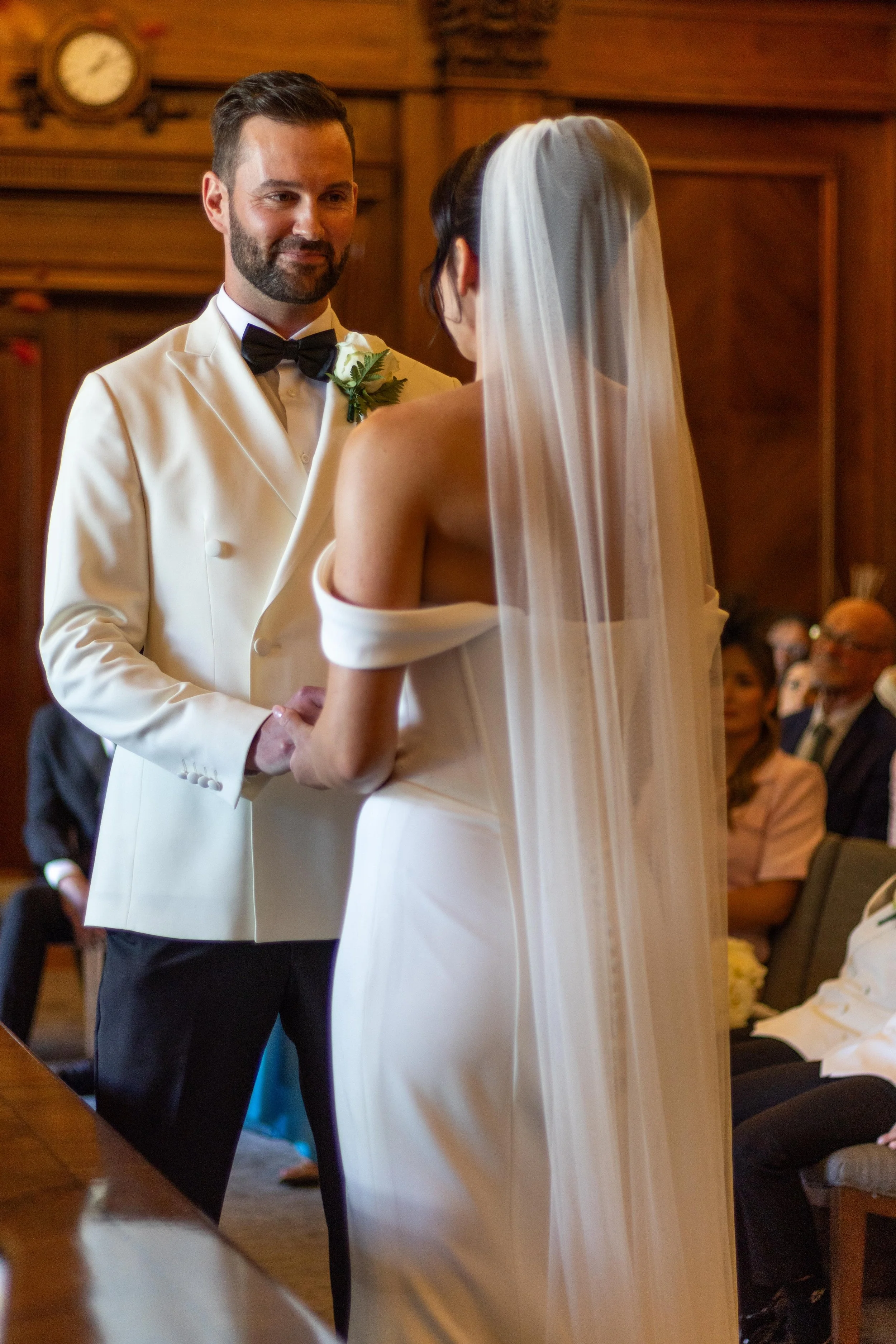 A bride and groom exchange vows during a wedding ceremony in a wood-paneled room. The groom is wearing a white tuxedo with a black bow tie, while the bride is in a white off-shoulder dress with a veil. Guests are seated in the background.