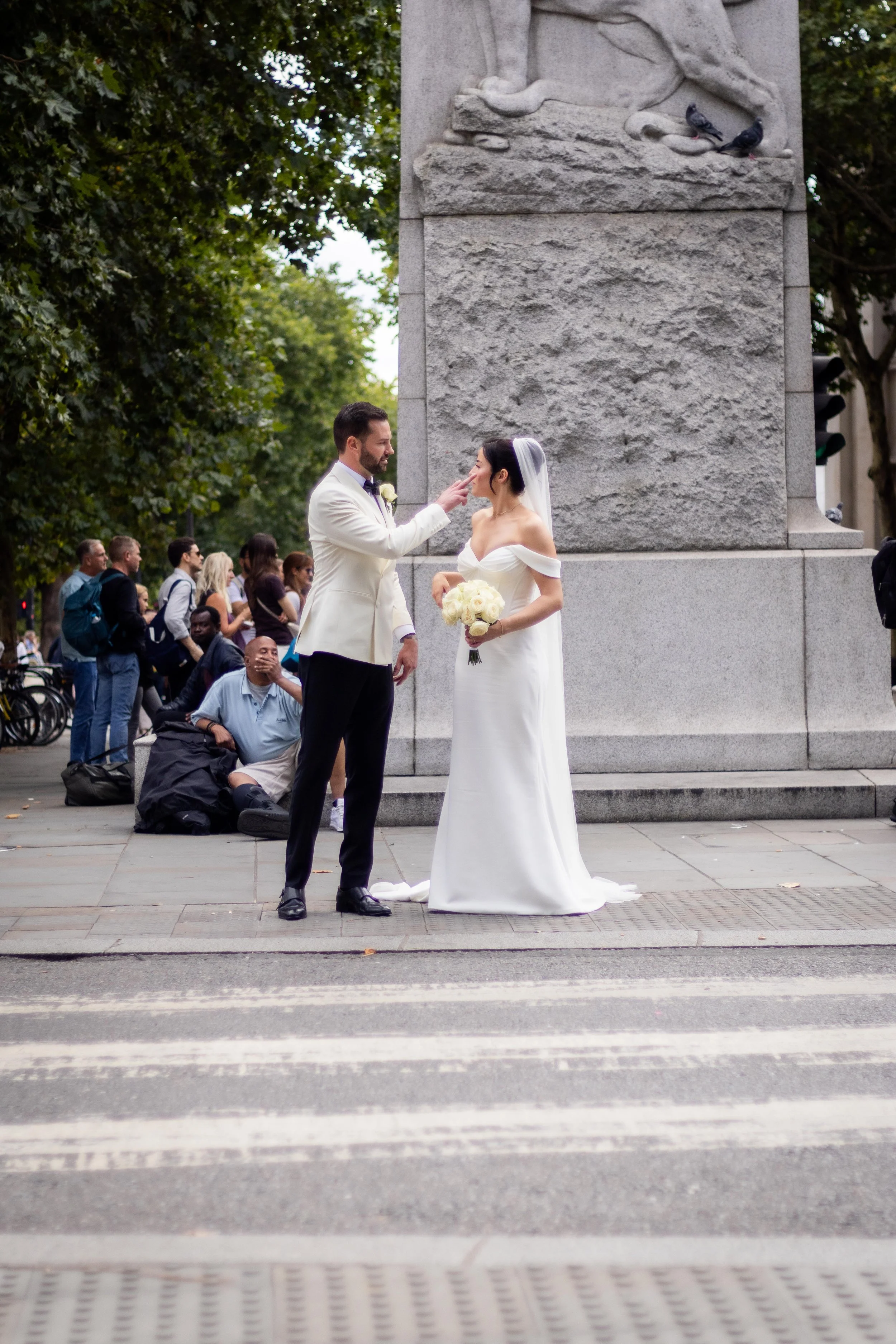 A bride and groom have a wedding ceremony on the street with an audience of onlookers in the background. The groom is touching the bride's face, who is holding a bouquet of white flowers, as they stand in front of a stone monument with carved sculptu