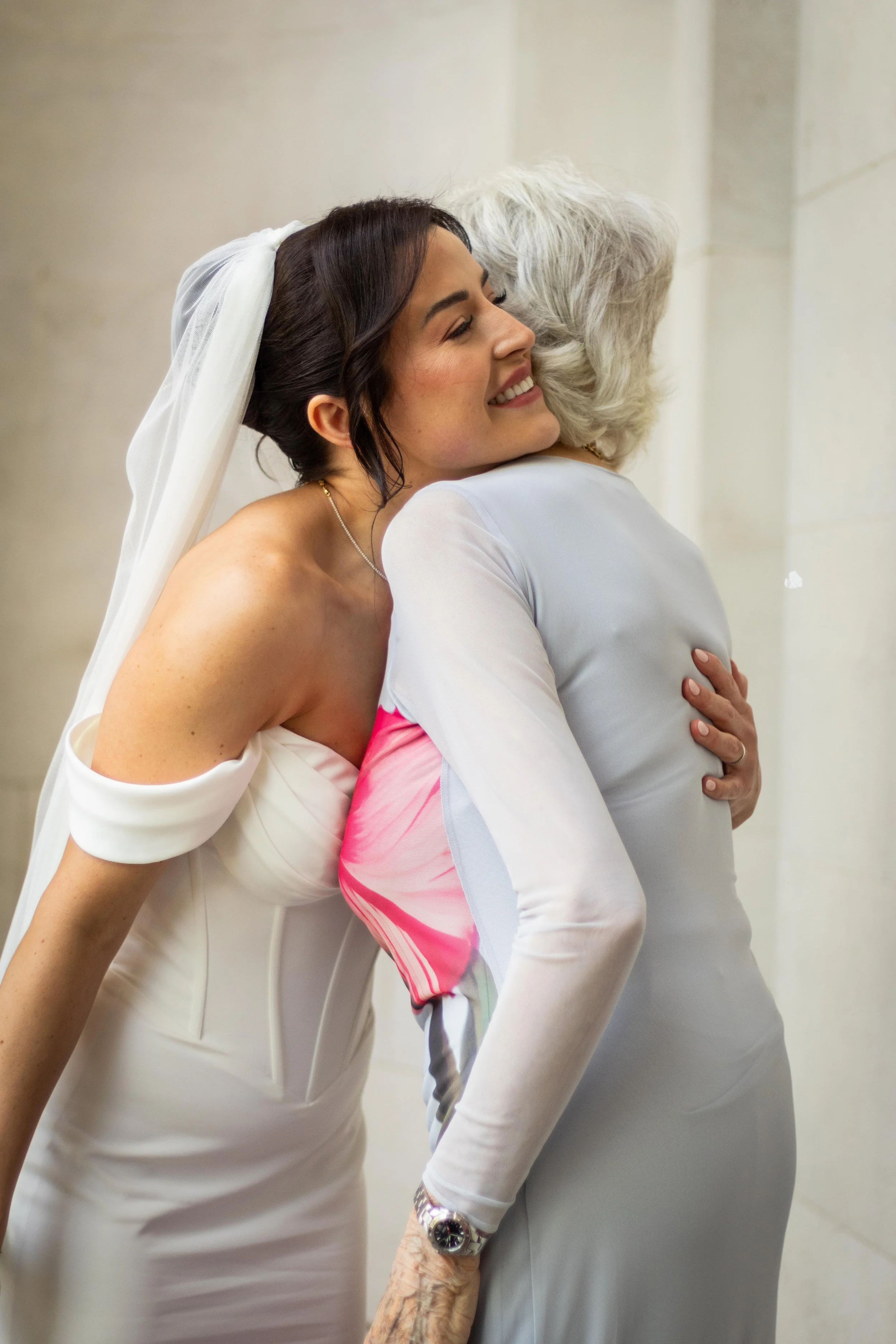 A young woman in a wedding dress hugging an older woman in a light-colored outfit, both smiling with their eyes closed.