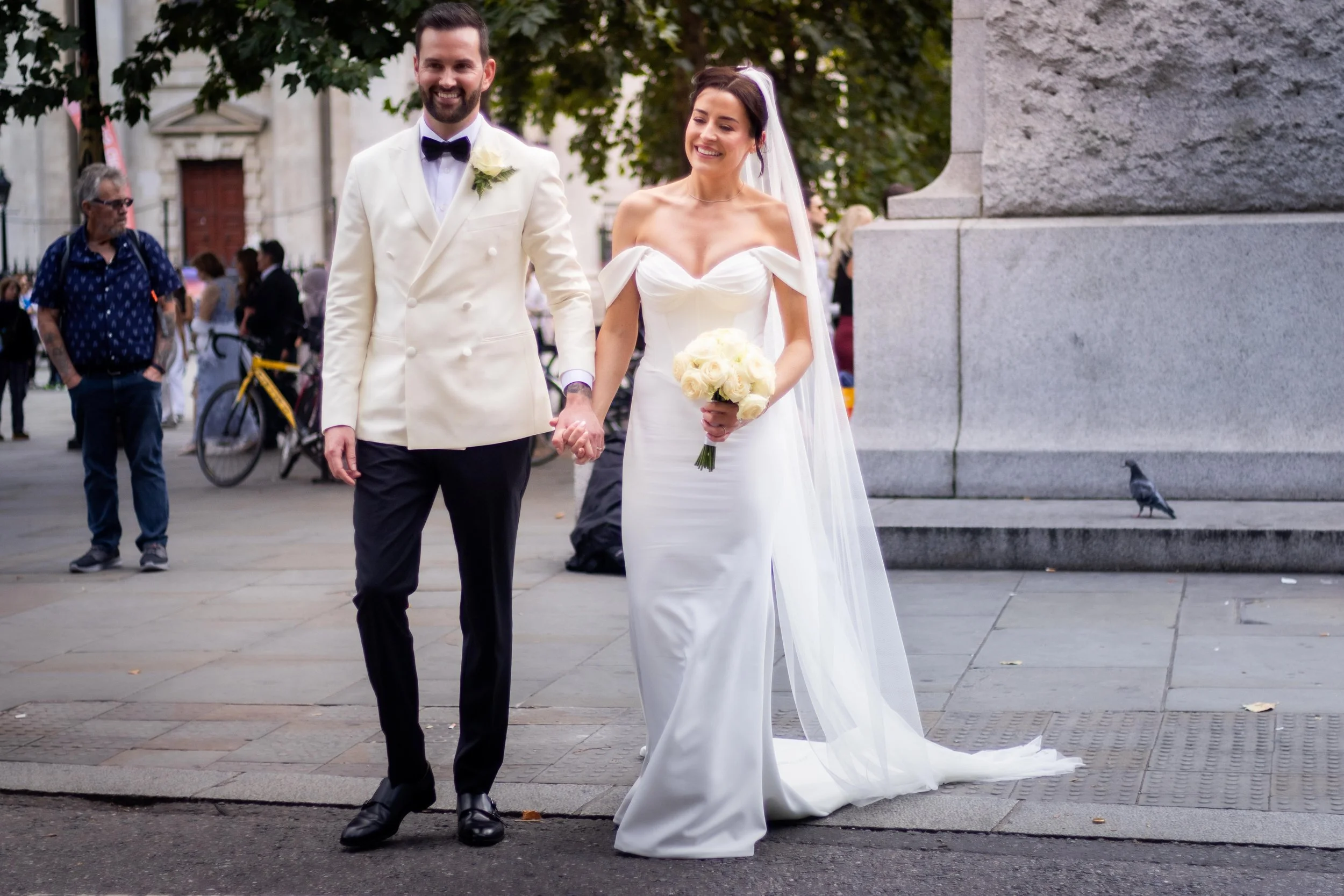 A newly married couple walking hand in hand outdoors. The groom is wearing a white tuxedo jacket with black pants, and the bride is dressed in a white wedding gown holding a bouquet of white roses.