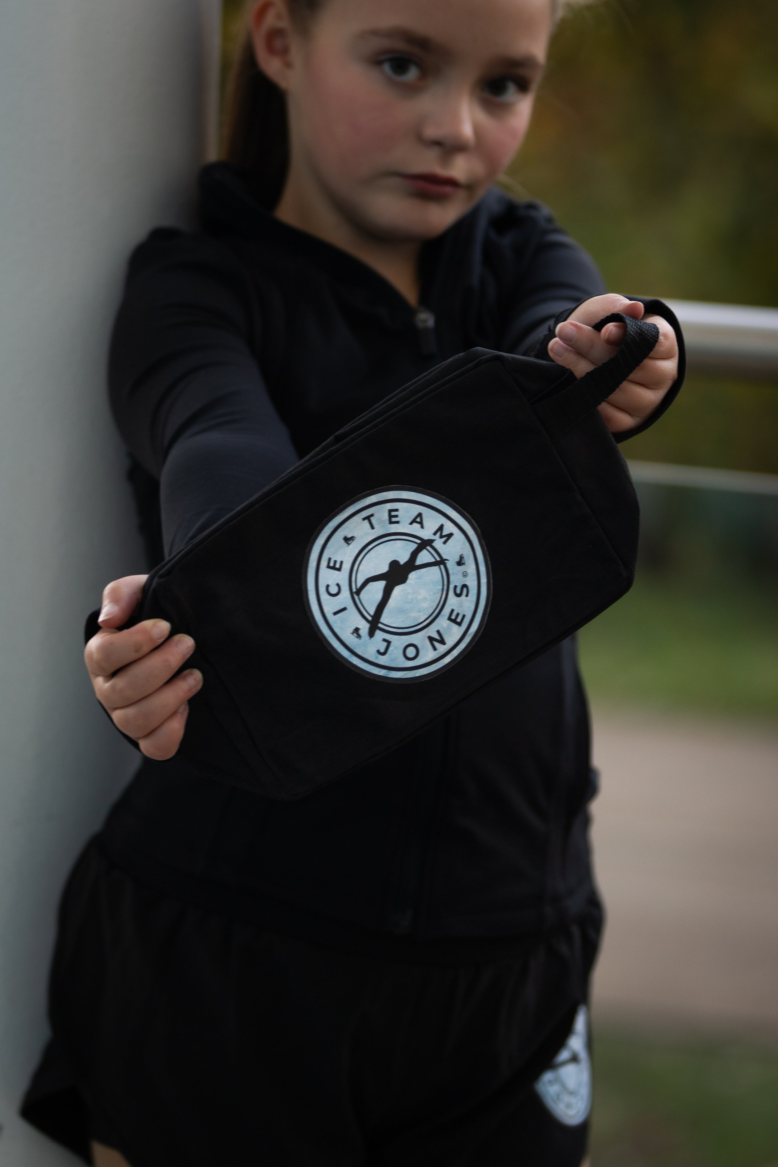A young girl holding a black bag with the Ice Team Jones logo, standing outdoors near a wall, wearing a black jacket.
