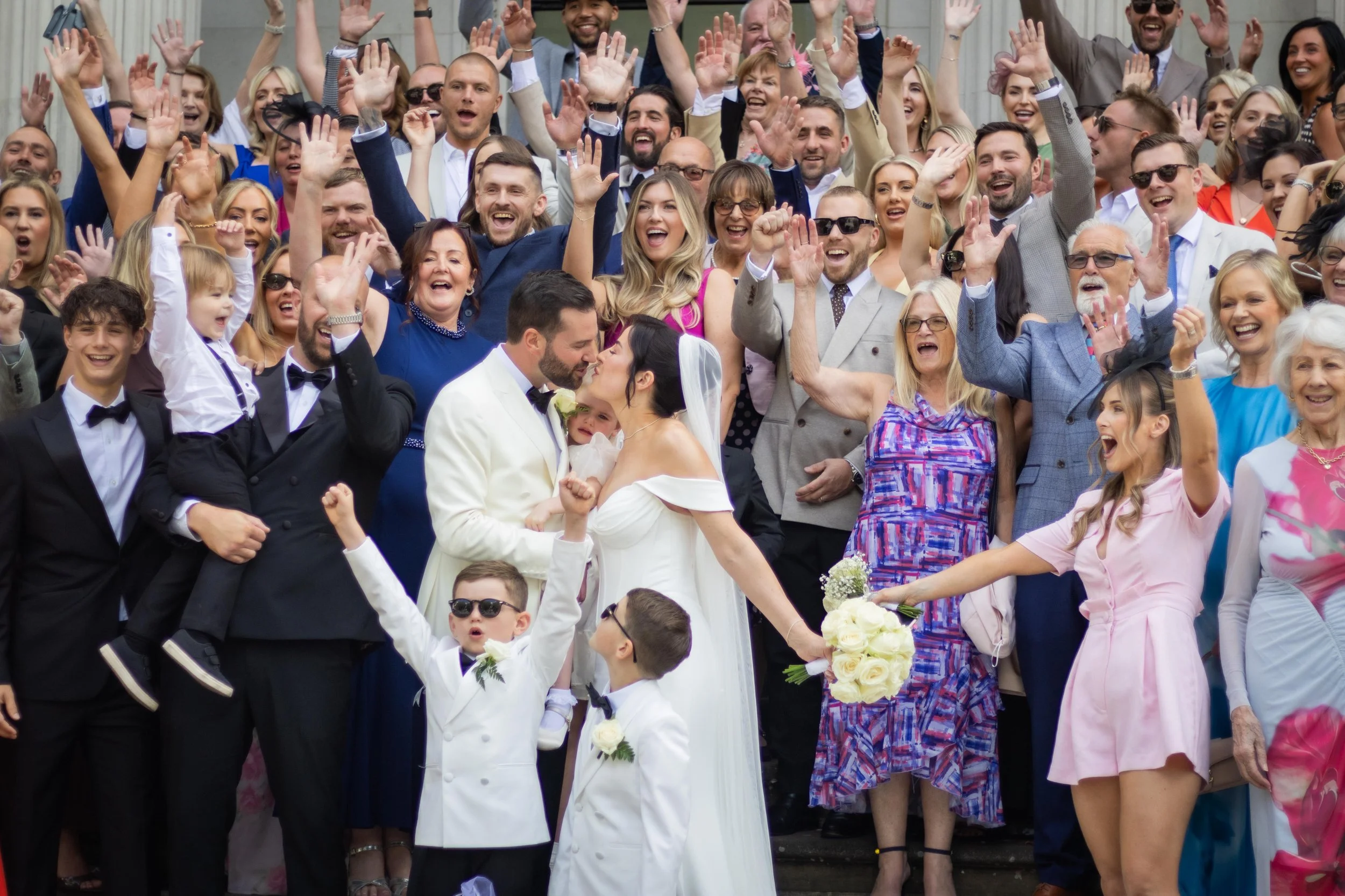 A large group of people gathered outside celebrating a wedding, with the bride and groom kissing in the center. The bride is holding a bouquet of white roses, and children and adults are surrounding them, dressed in formal and colorful attire, smilin