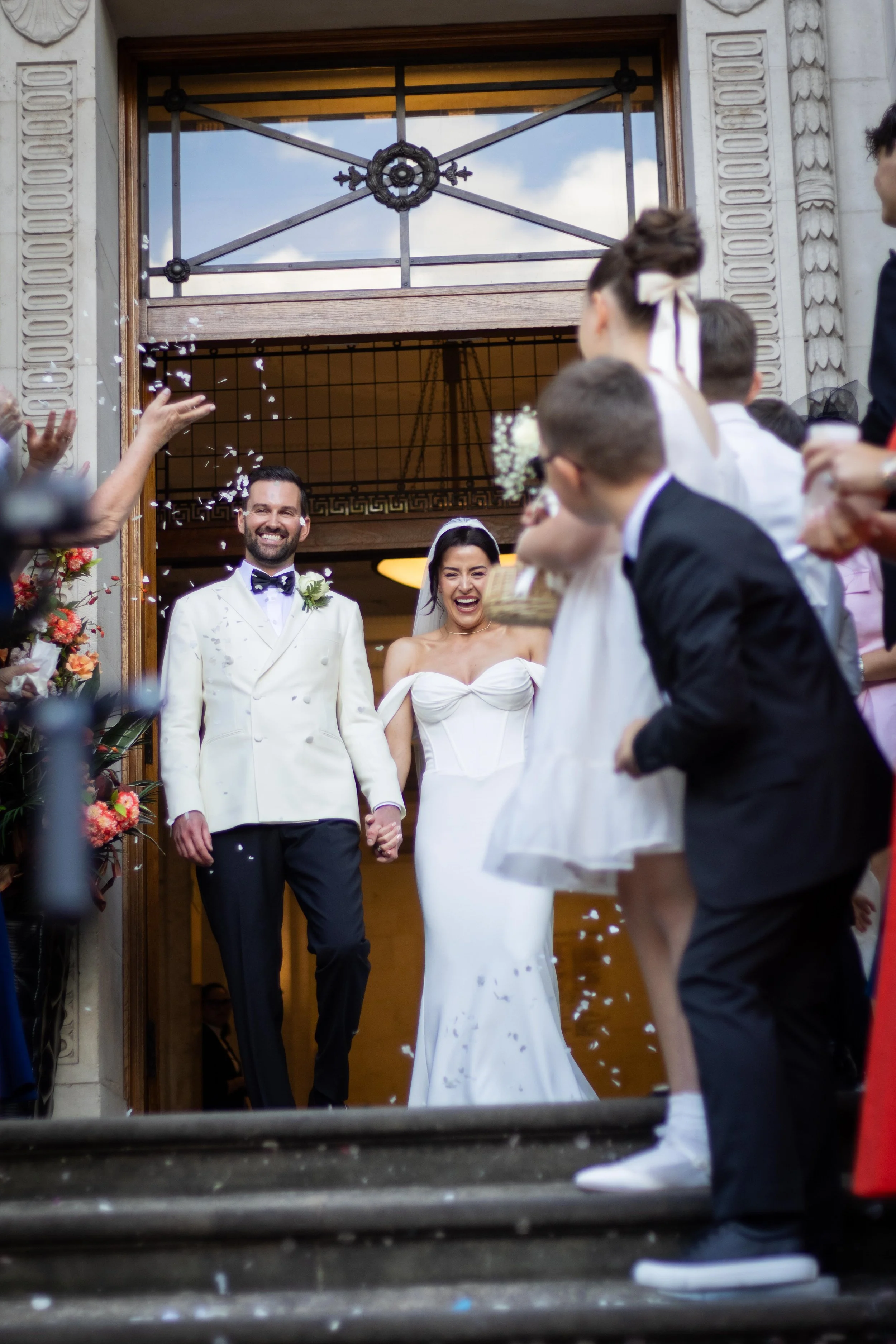 A newlywed couple holding hands, smiling as they walk down the stairs surrounded by friends and family celebrating with confetti outside a building.
