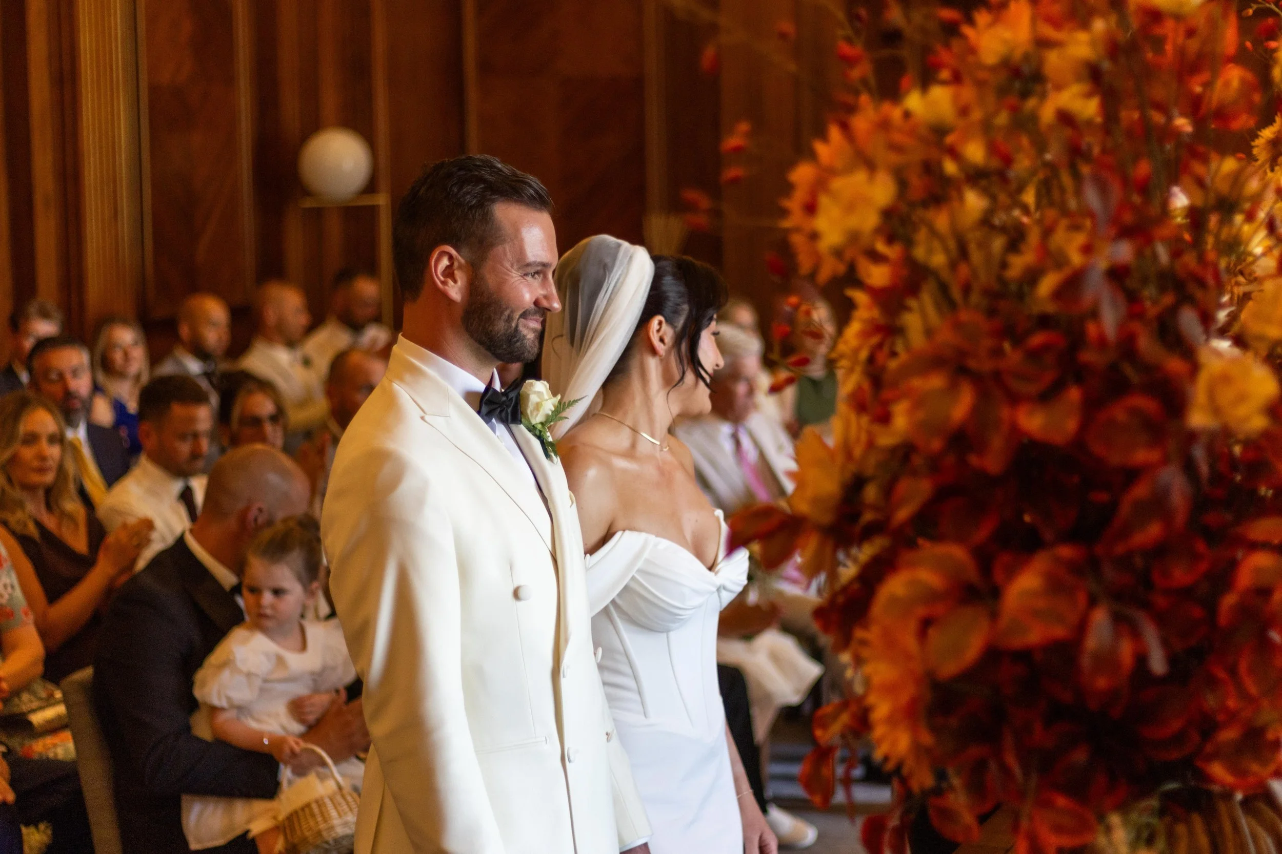 A bride and groom standing at the altar during their wedding ceremony, surrounded by seated guests, with a large floral arrangement in the foreground.