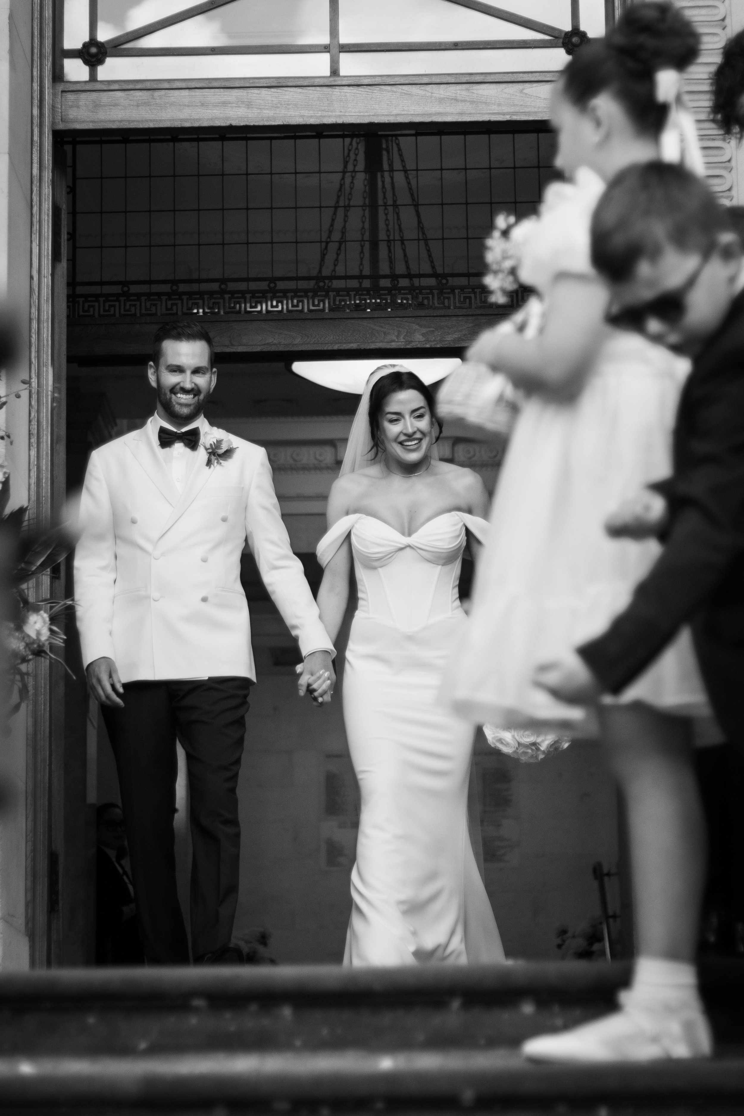 Black and white photo of a newlywed couple holding hands and smiling, walking down the aisle inside a church, surrounded by children.