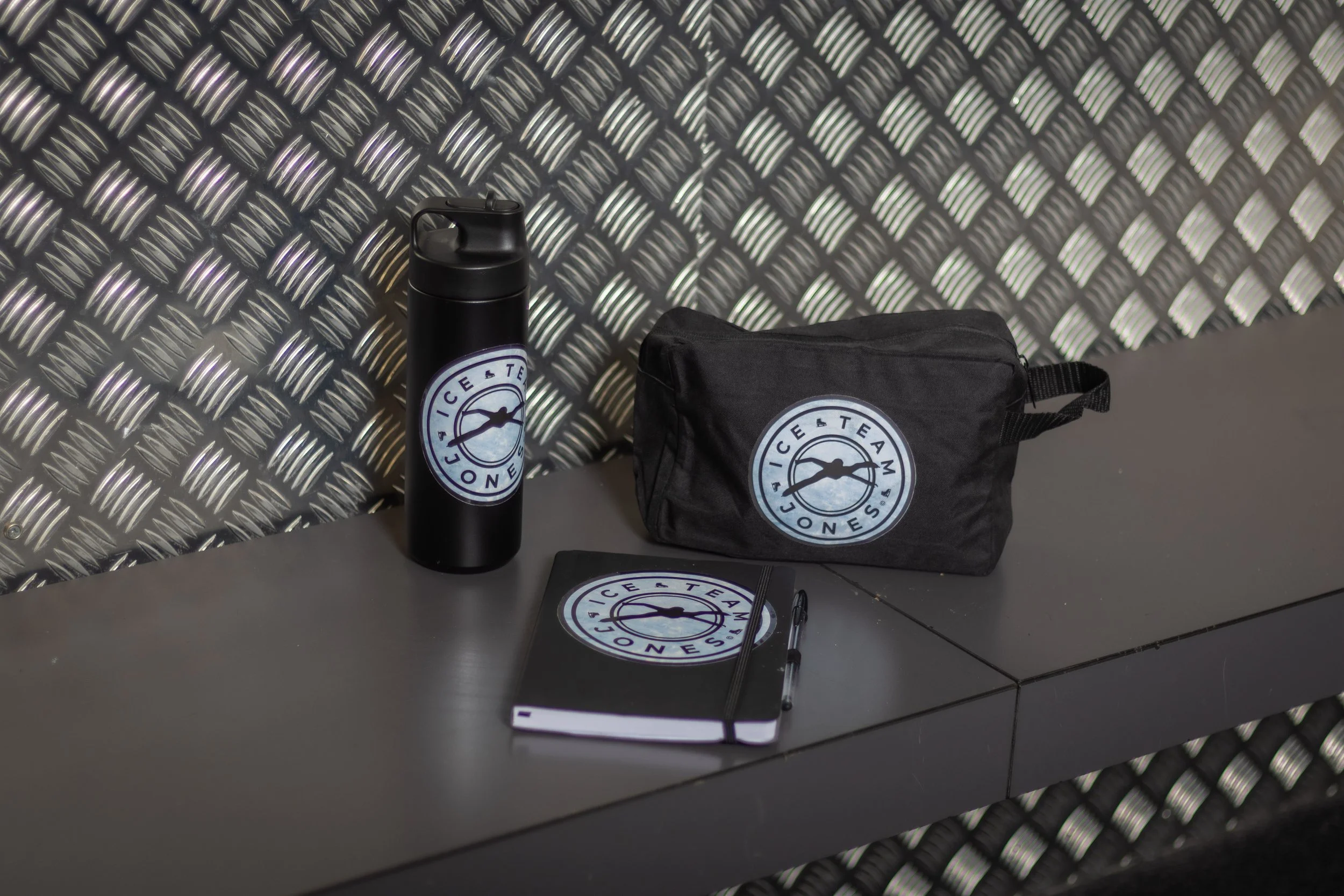A black water bottle, a black zippered pouch, a notebook with a logo, and a pen with a logo on a gray tabletop against a textured metallic wall.