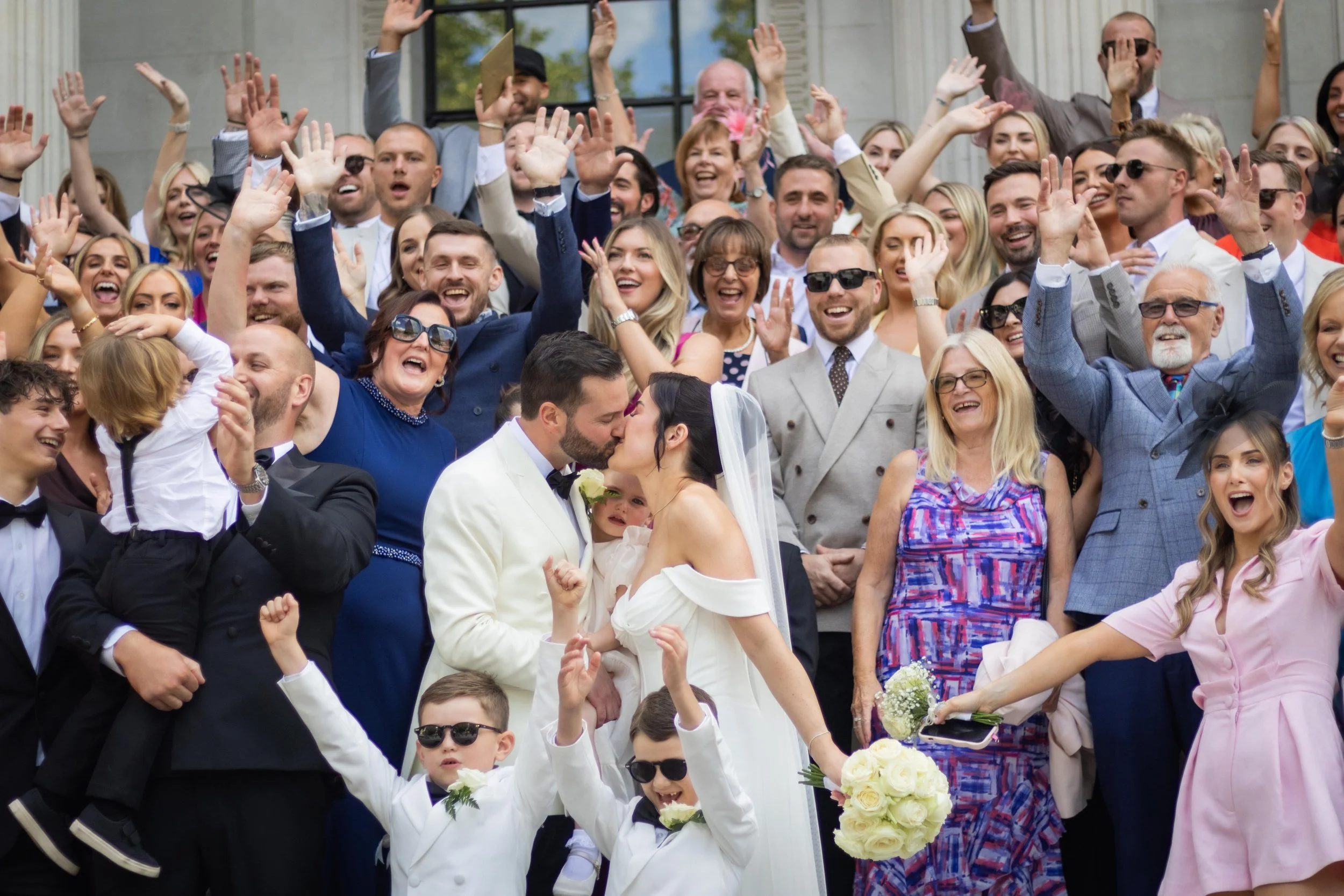 A large group of smiling people celebrating at a wedding, with the bride and groom kissing in the center.
