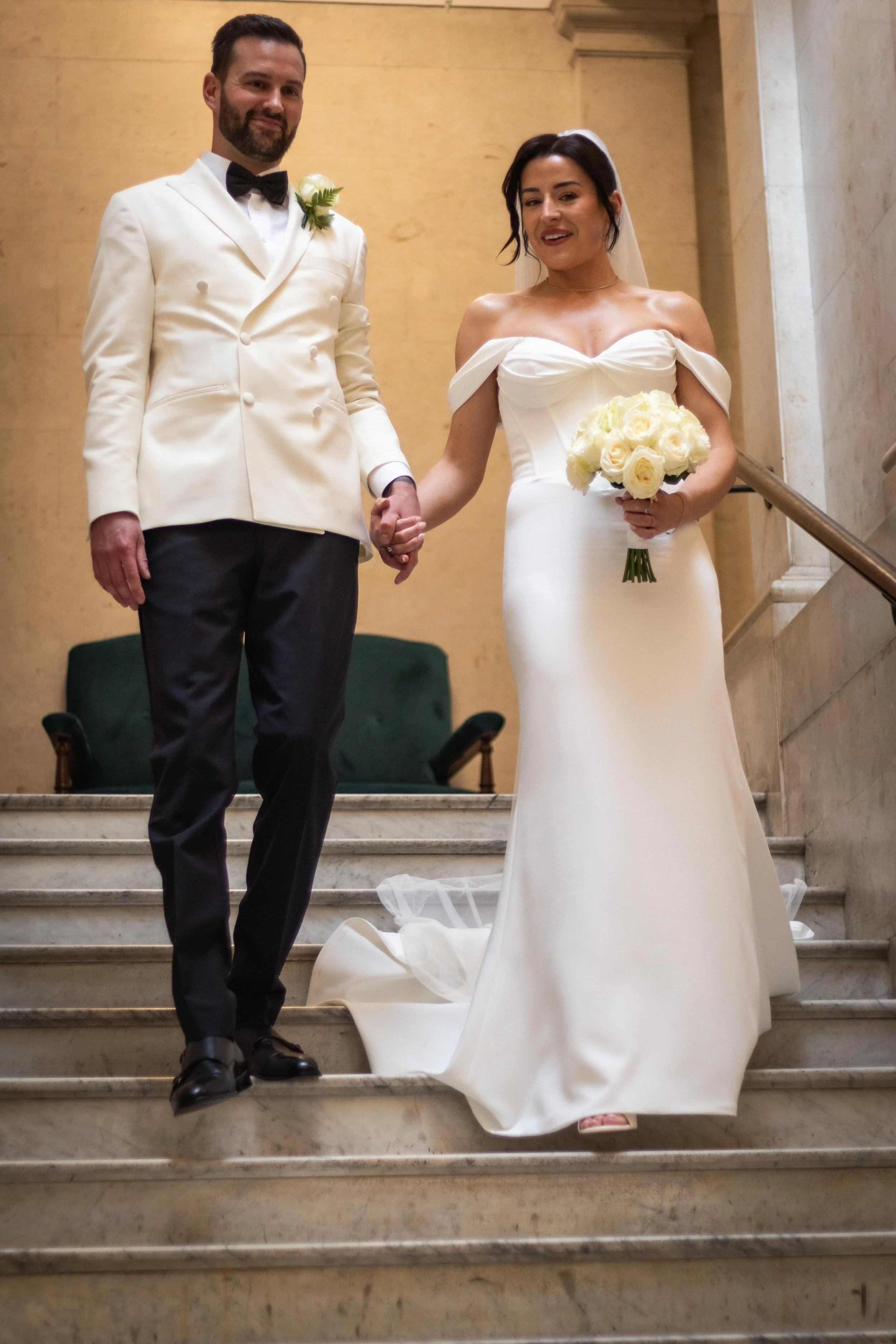 A newlywed couple standing on stairs inside a building, holding hands. The groom is dressed in a white tuxedo jacket with a black bow tie and black pants. The bride is wearing a white off-shoulder wedding dress and holding a bouquet of white roses. T