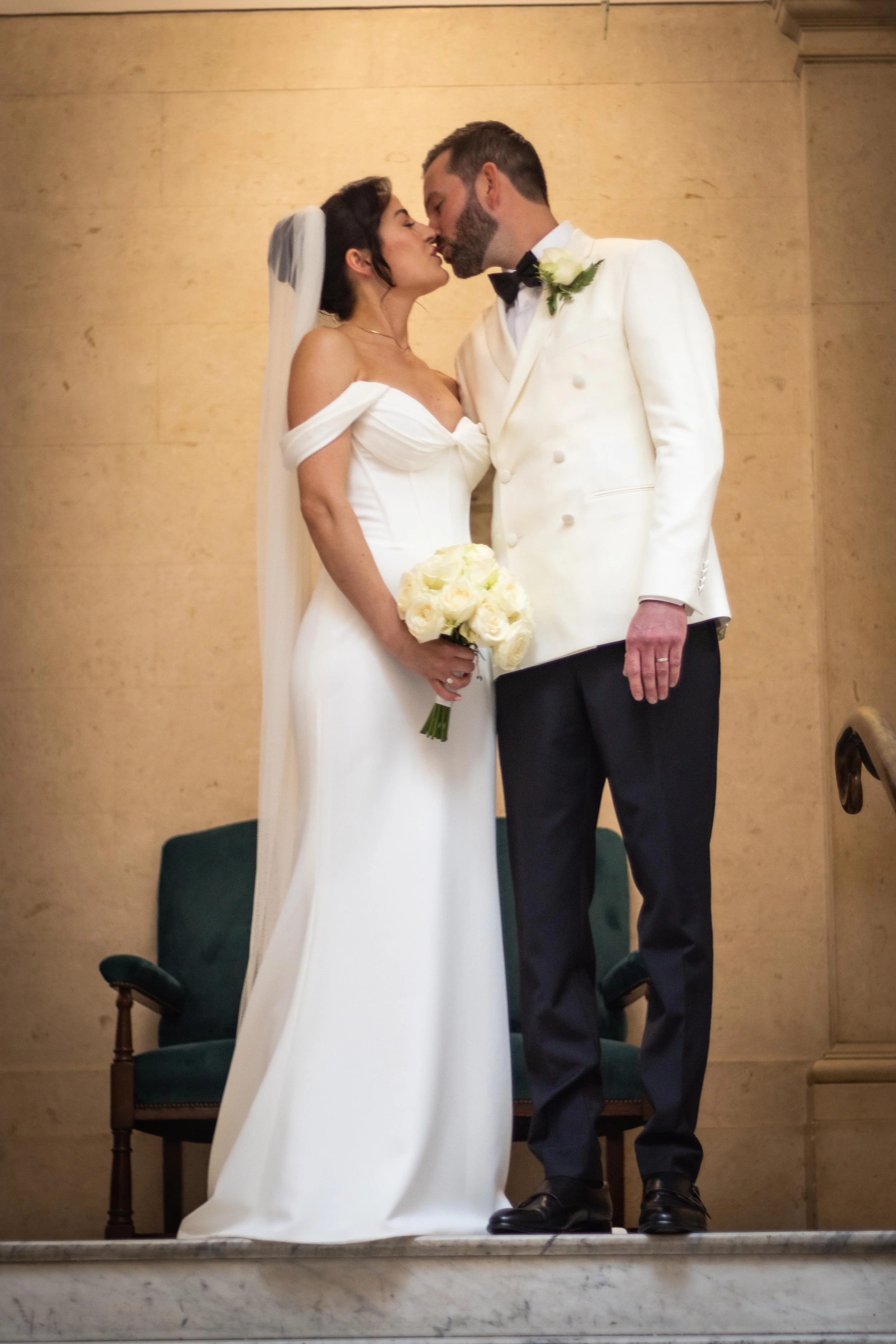 A bride and groom share a kiss during their wedding ceremony, with the bride holding a bouquet of white roses, both dressed in formal wedding attire.