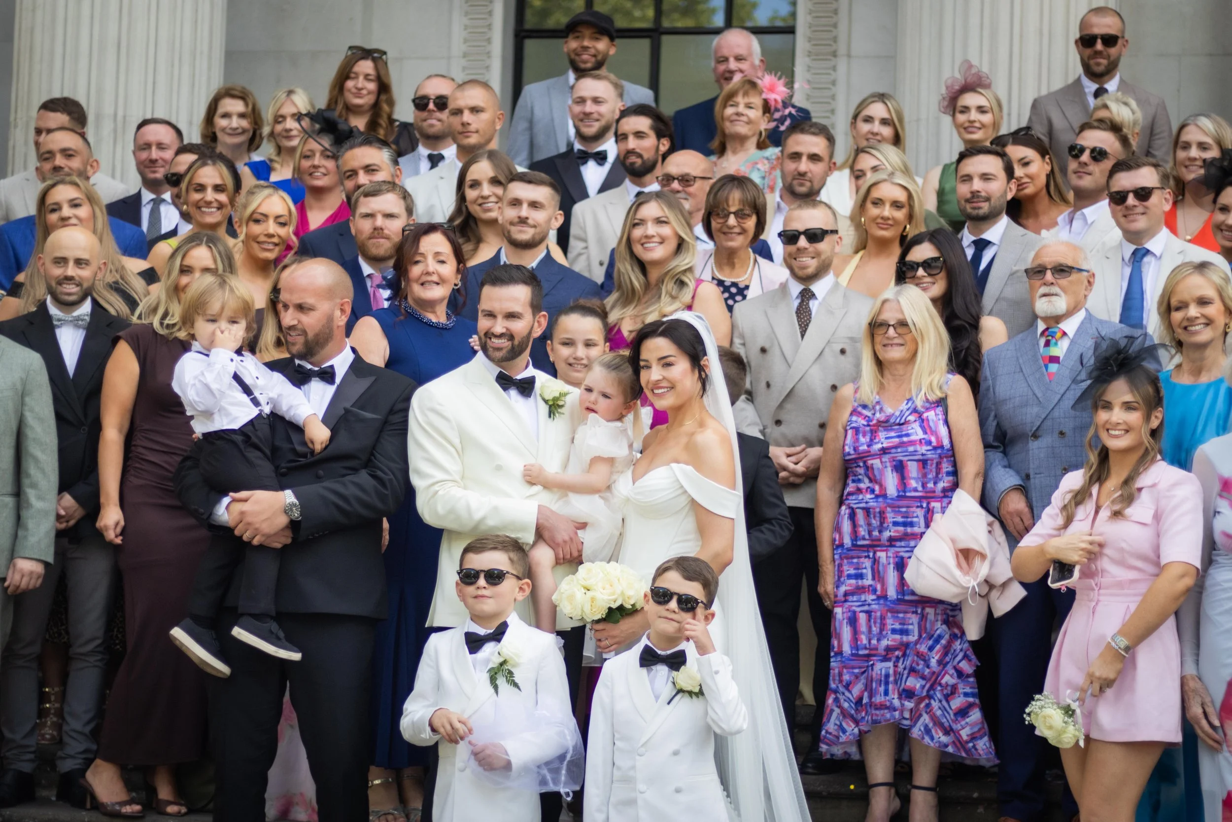 Group of wedding guests on the steps of a building, including a bride and groom in the center, surrounded by family and friends.