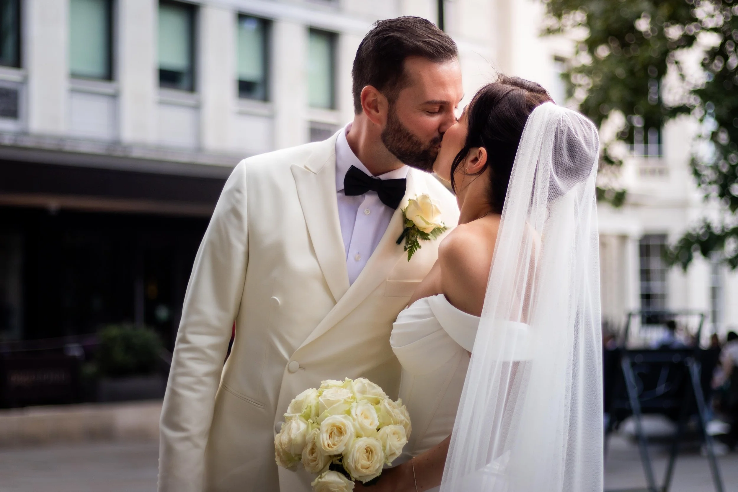 A bride and groom kiss outdoors at their wedding. The groom wears a white tuxedo with a black bow tie, and the bride is in a white gown with a veil, holding a bouquet of white roses.