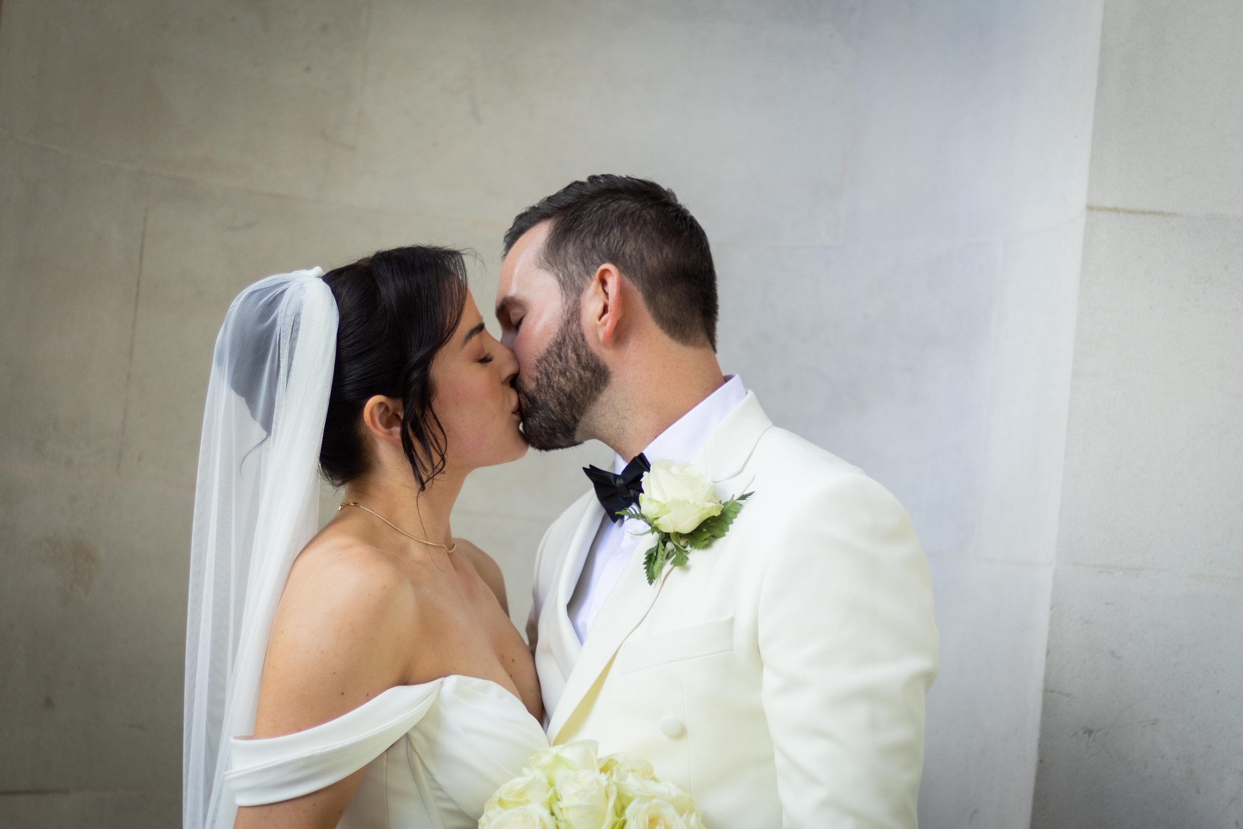 A bride and groom kissing, dressed in wedding attire against a plain light-colored wall.