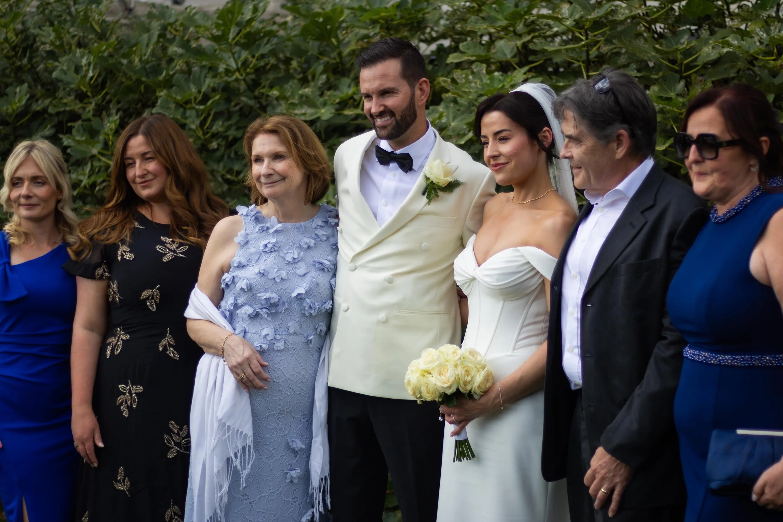Group of people at a wedding, including the bride in a white dress holding a bouquet of white roses, the groom in a white tuxedo, and other guests dressed in formal attire, standing outdoors with greenery in the background.