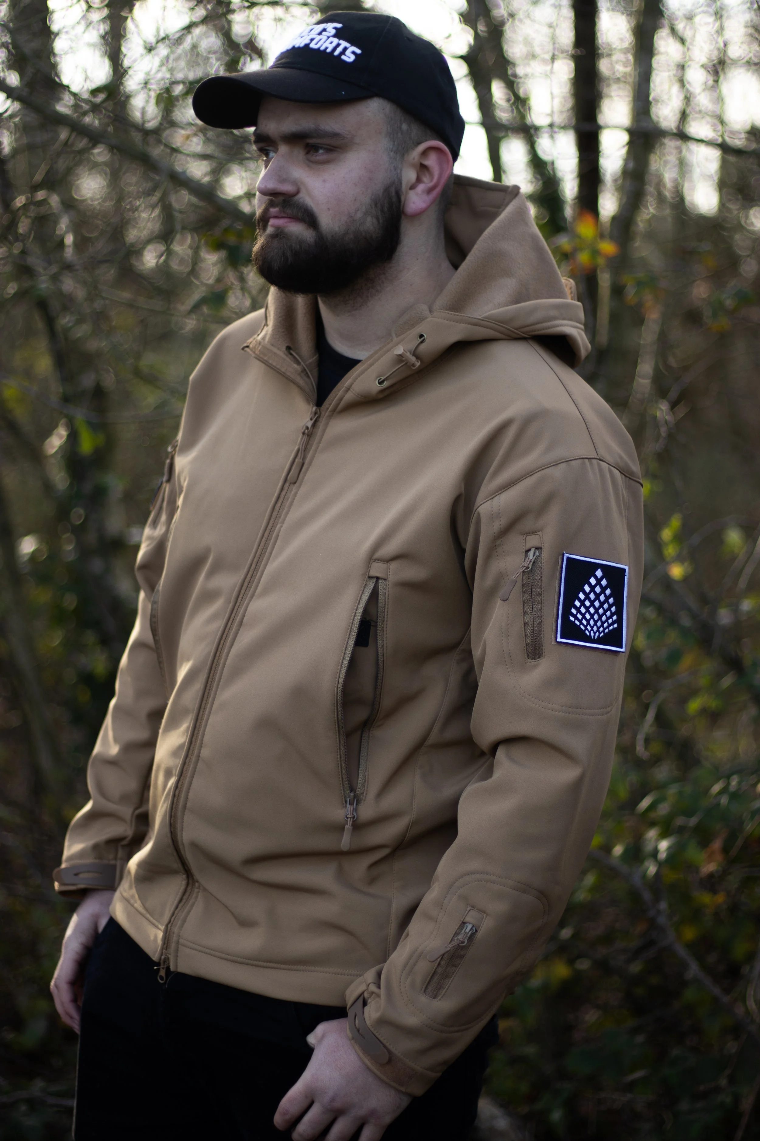 A man wearing a tan tactical jacket with a patch on the sleeve, standing outdoors in a wooded area with trees and branches in the background.