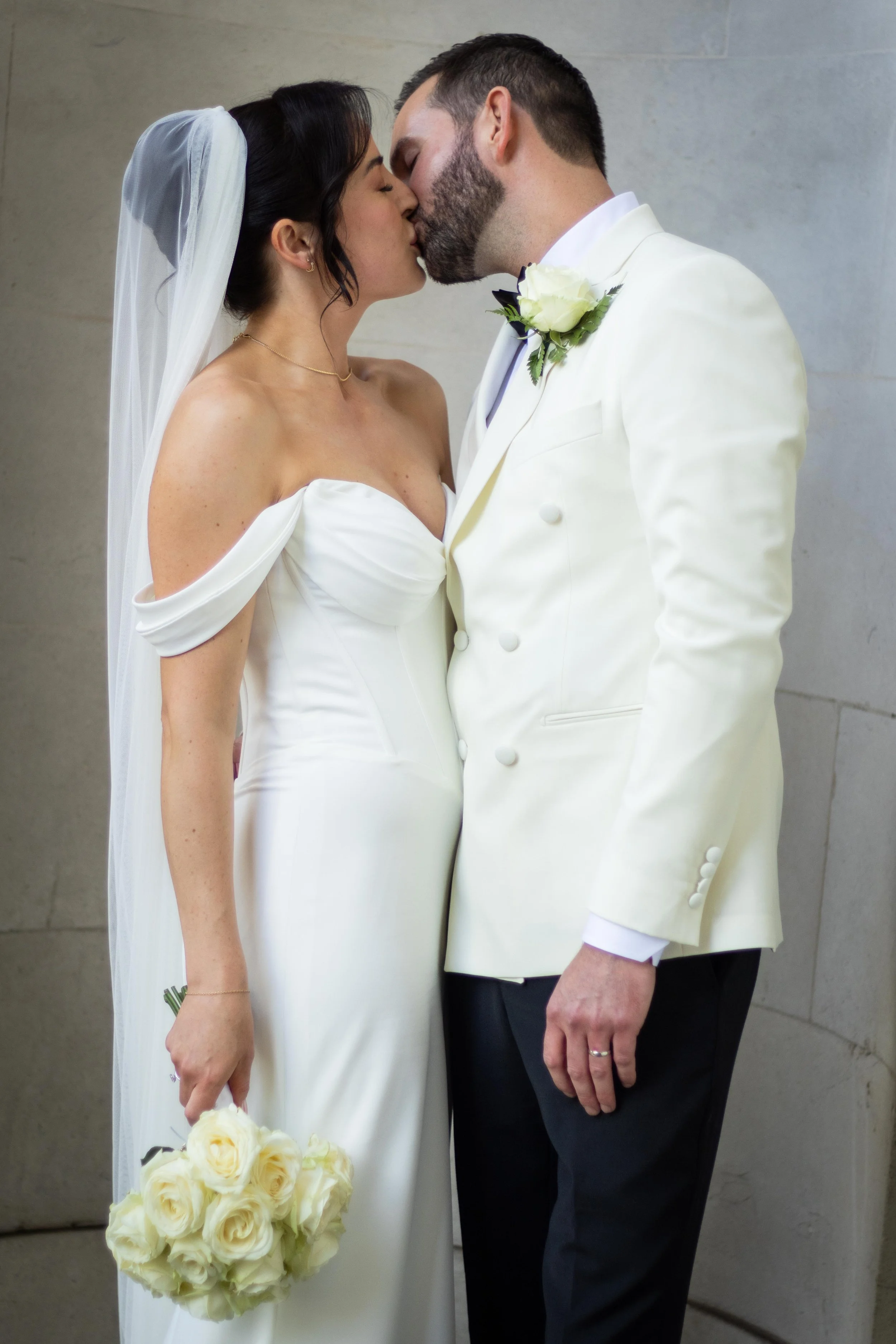 A bride and groom kissing at their wedding, the bride holding a bouquet of white roses, the groom wearing a cream-colored tuxedo with a white rose boutonniere.