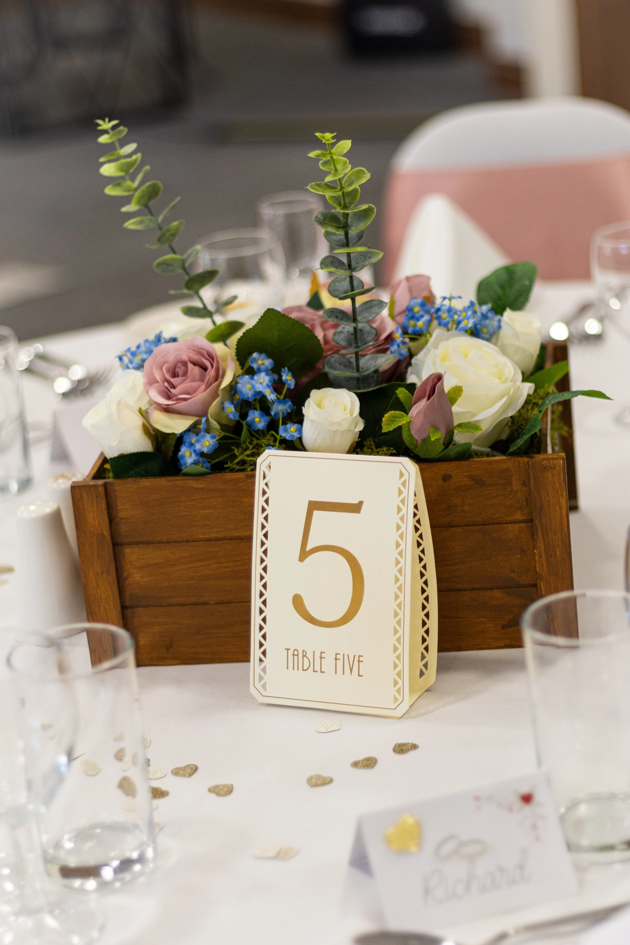 Table centerpiece with pink, white, and purple flowers in a wooden box, table number 5, and small heart-shaped confetti on a white tablecloth.