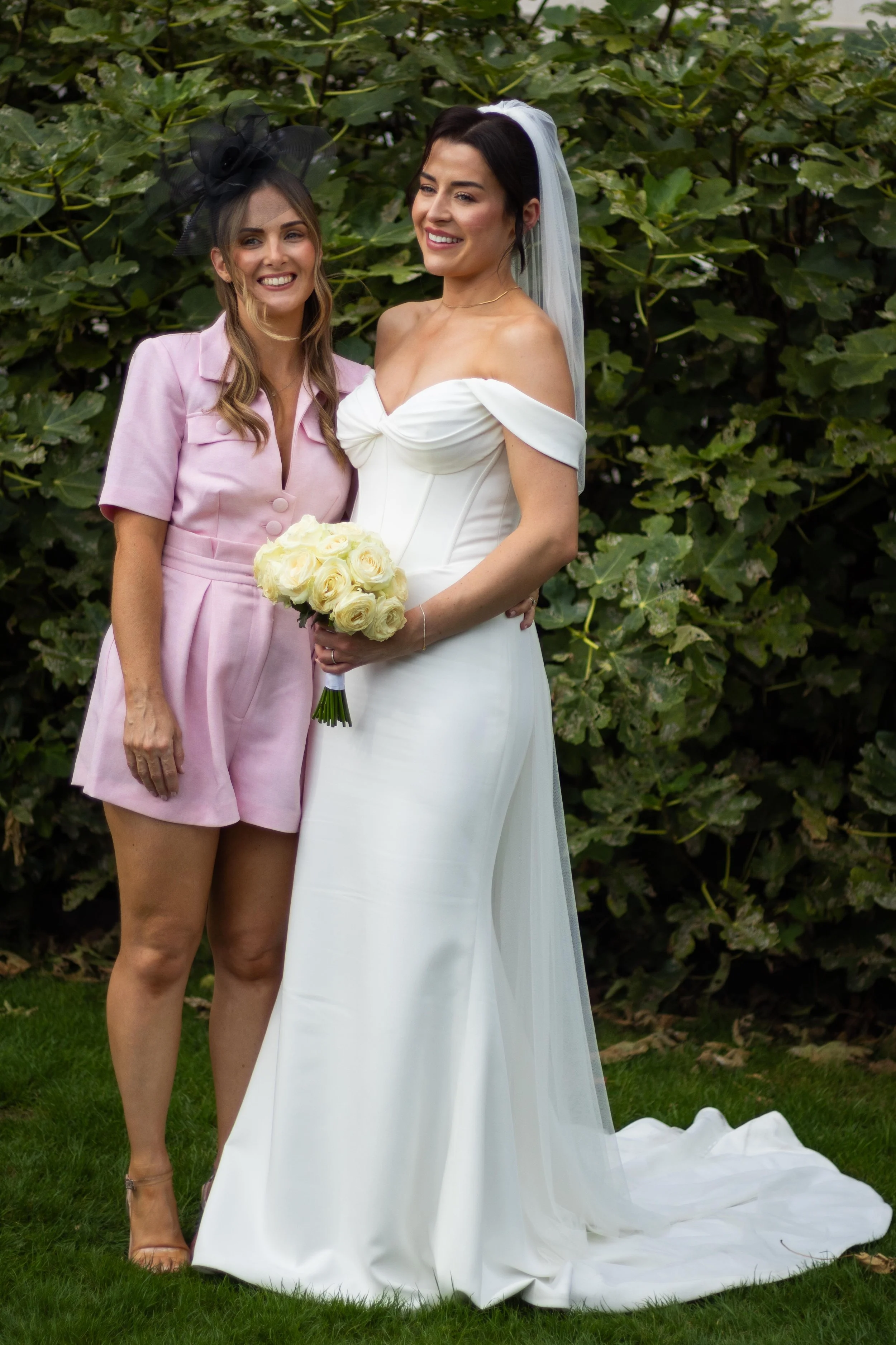 Two women, one in a wedding dress and veil holding a bouquet of white roses, and the other in a pink outfit with a black fascinator, standing outdoors in front of green foliage, smiling.
