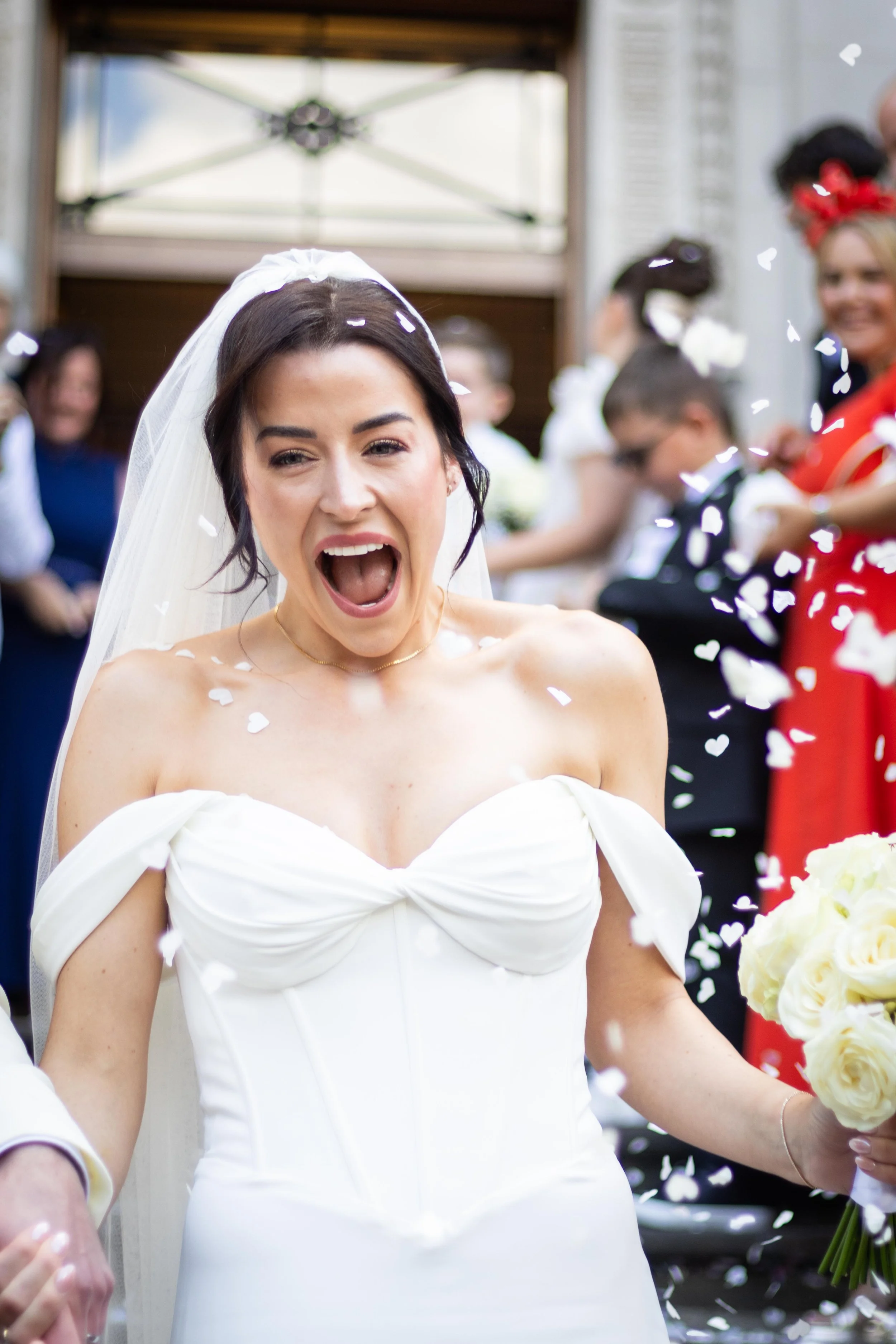 A bride with dark hair wearing a white wedding dress and veil, smiling and celebrating with confetti, holding a bouquet of white roses, surrounded by guests.
