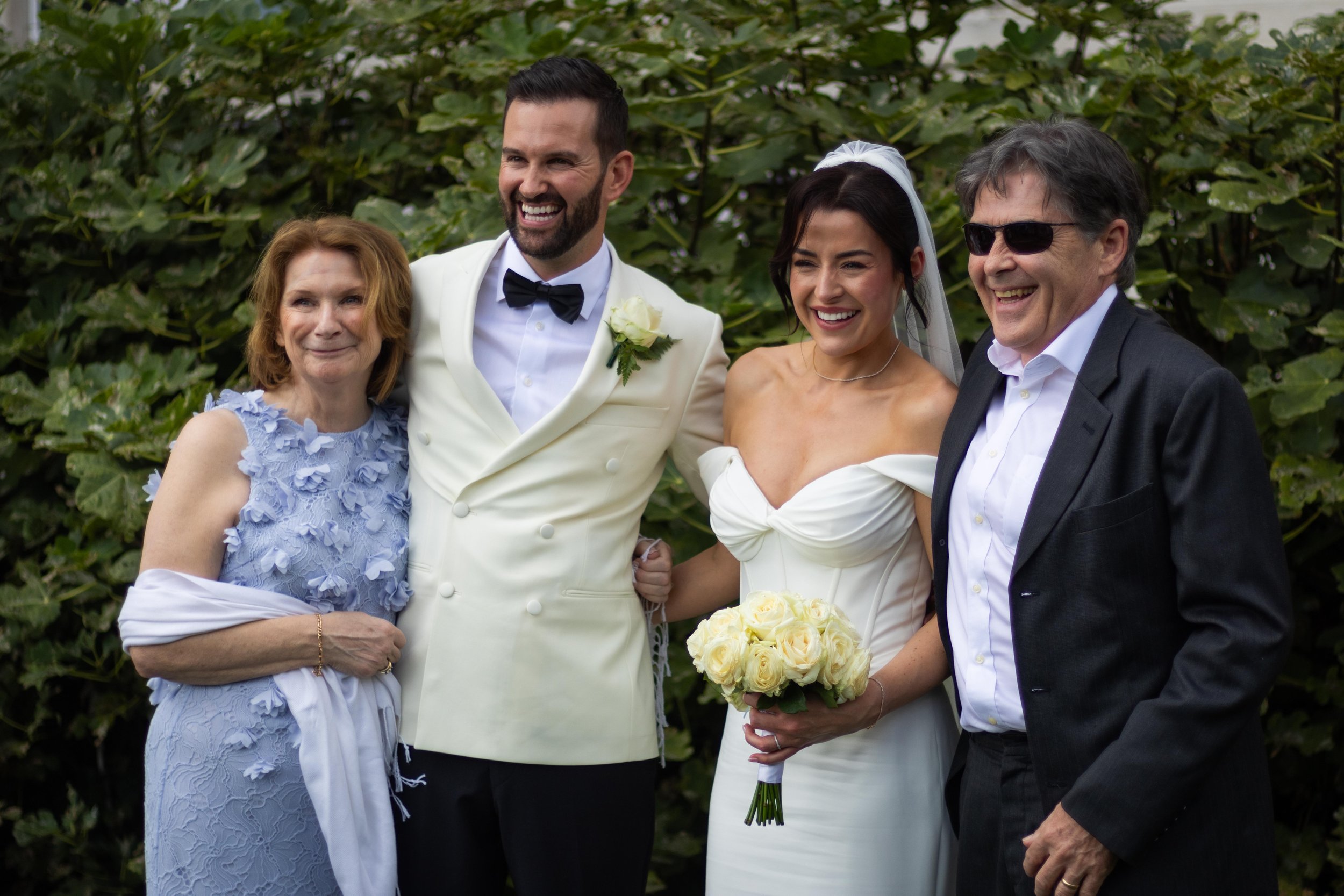 A group of four people standing outdoors, celebrating a wedding. The bride is holding a bouquet of white roses and is wearing a strapless white wedding dress, with a veil. The groom is beside her, dressed in a white suit with a black bow tie. An olde