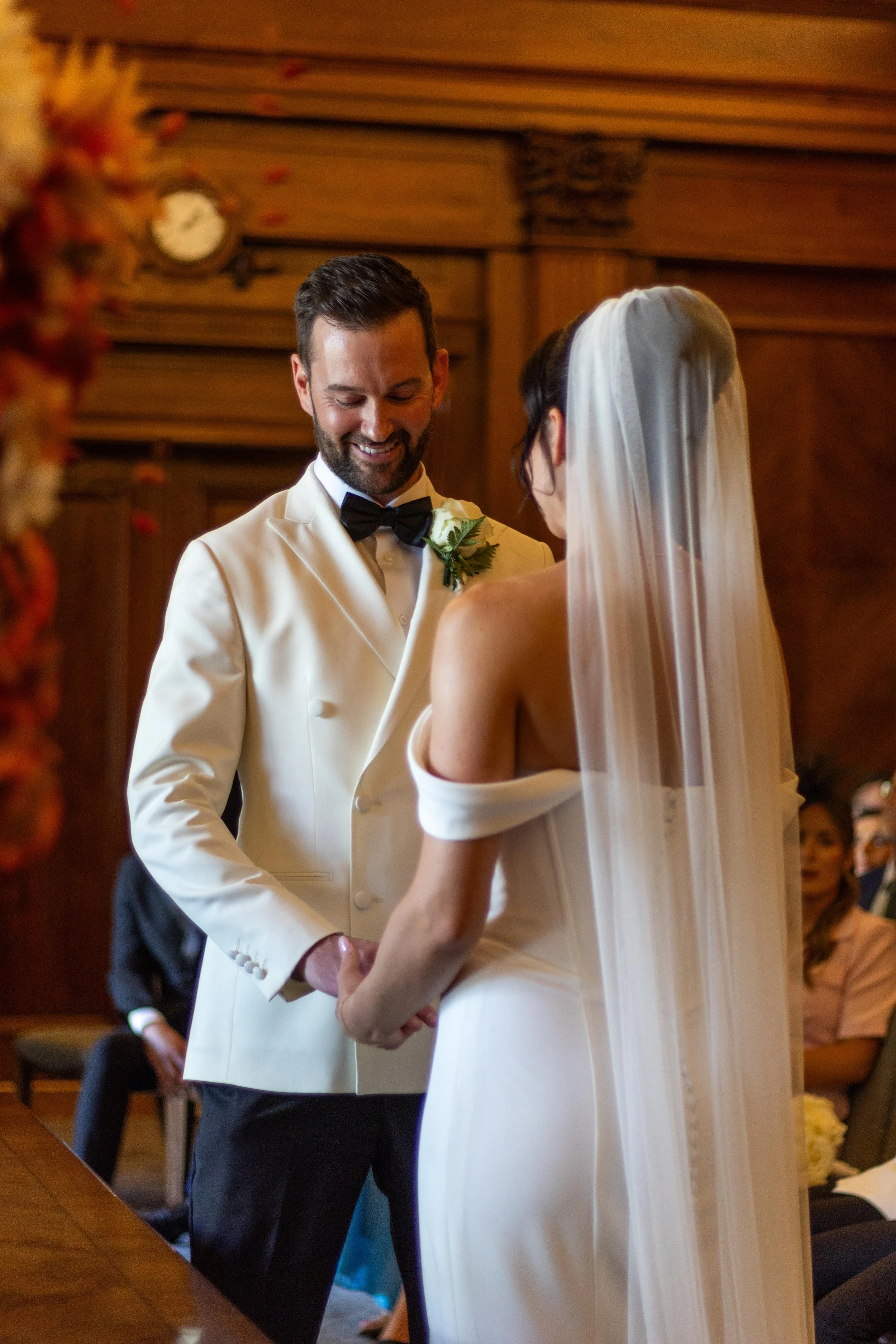 A bride and groom exchange vows in a wedding ceremony in a wood-paneled room, with other guests seated in the background.