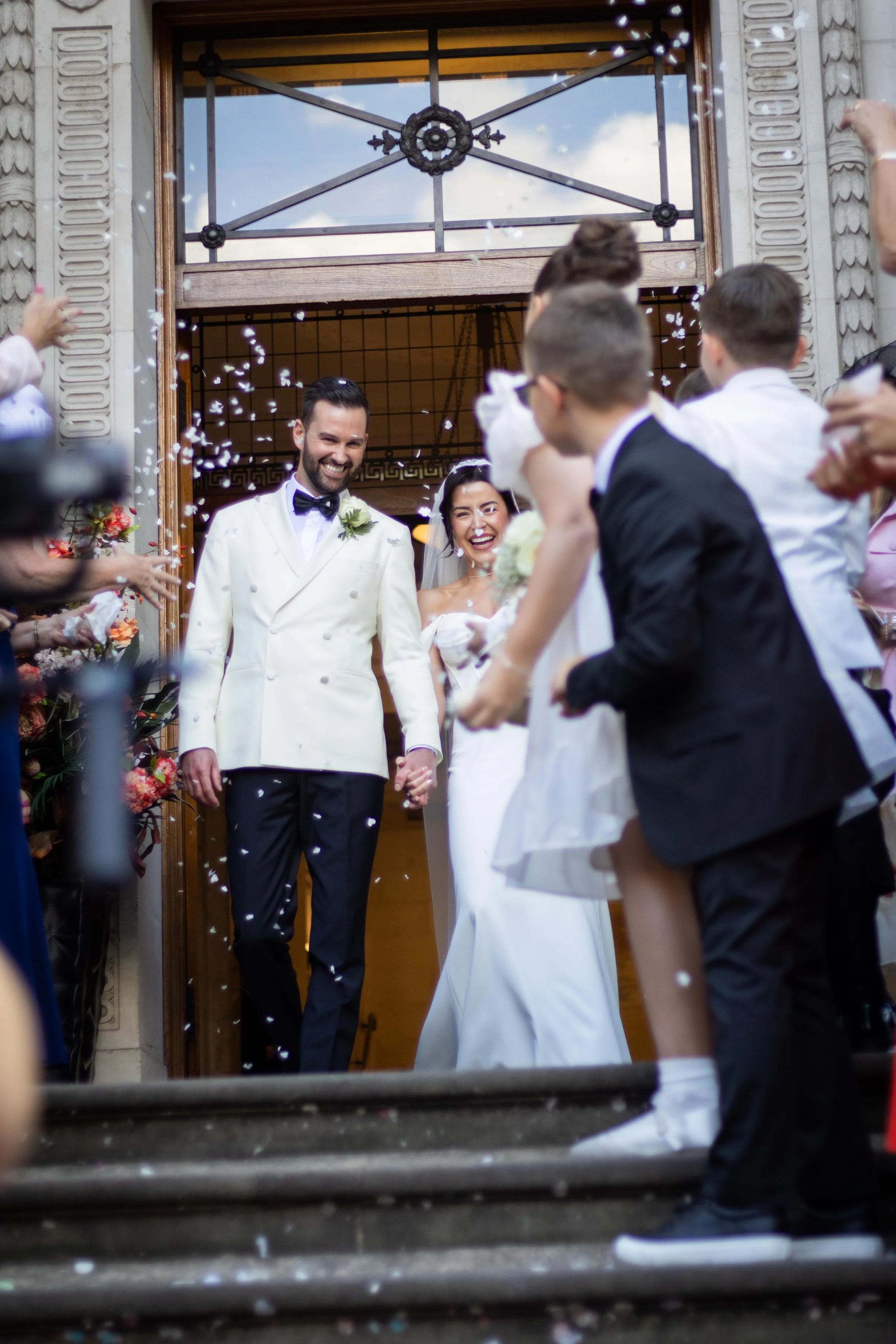 A newlywed couple exiting a building, holding hands and smiling, while being showered with confetti by friends and family.