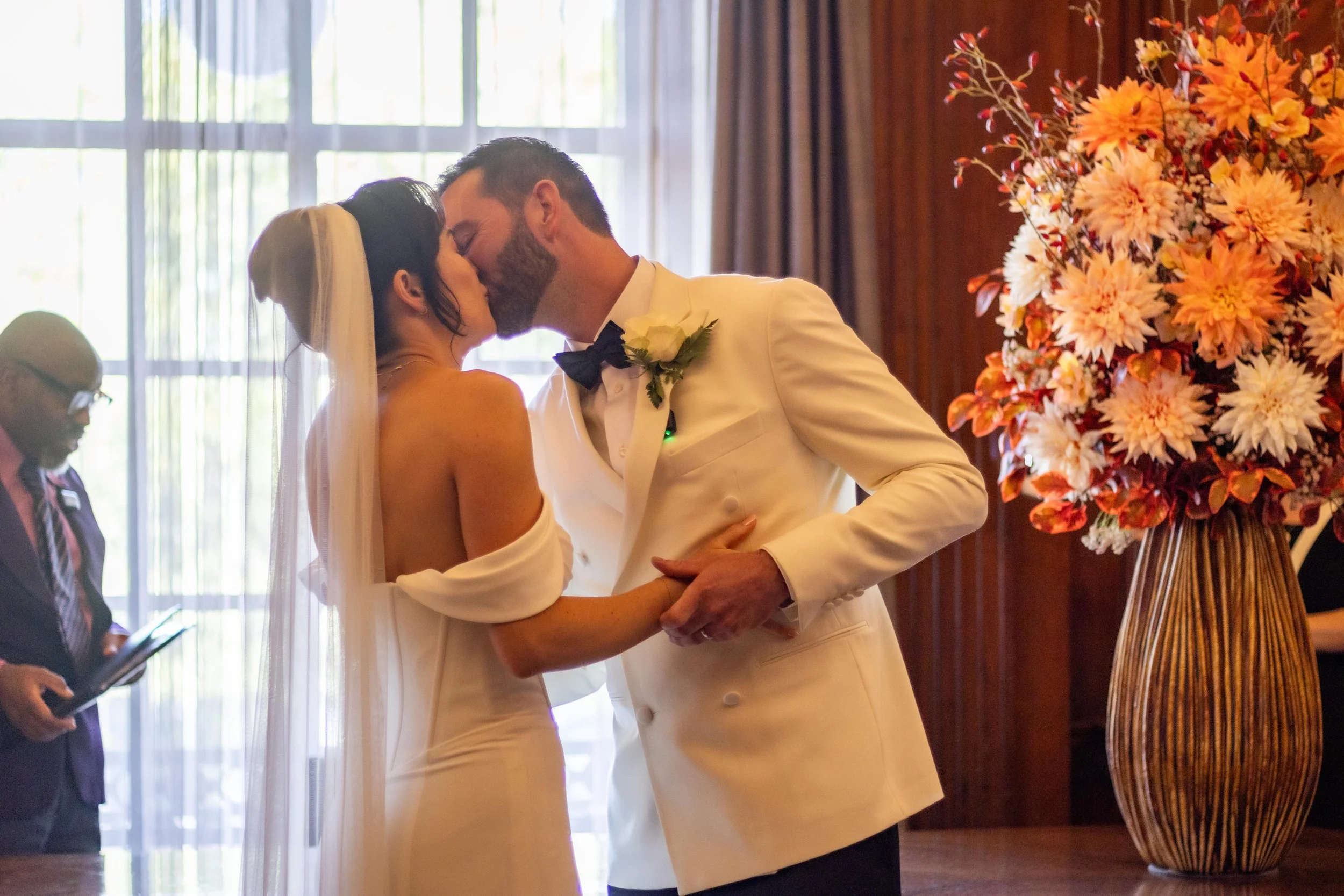 Bride and groom sharing a kiss during their wedding ceremony, with a large floral arrangement nearby.
