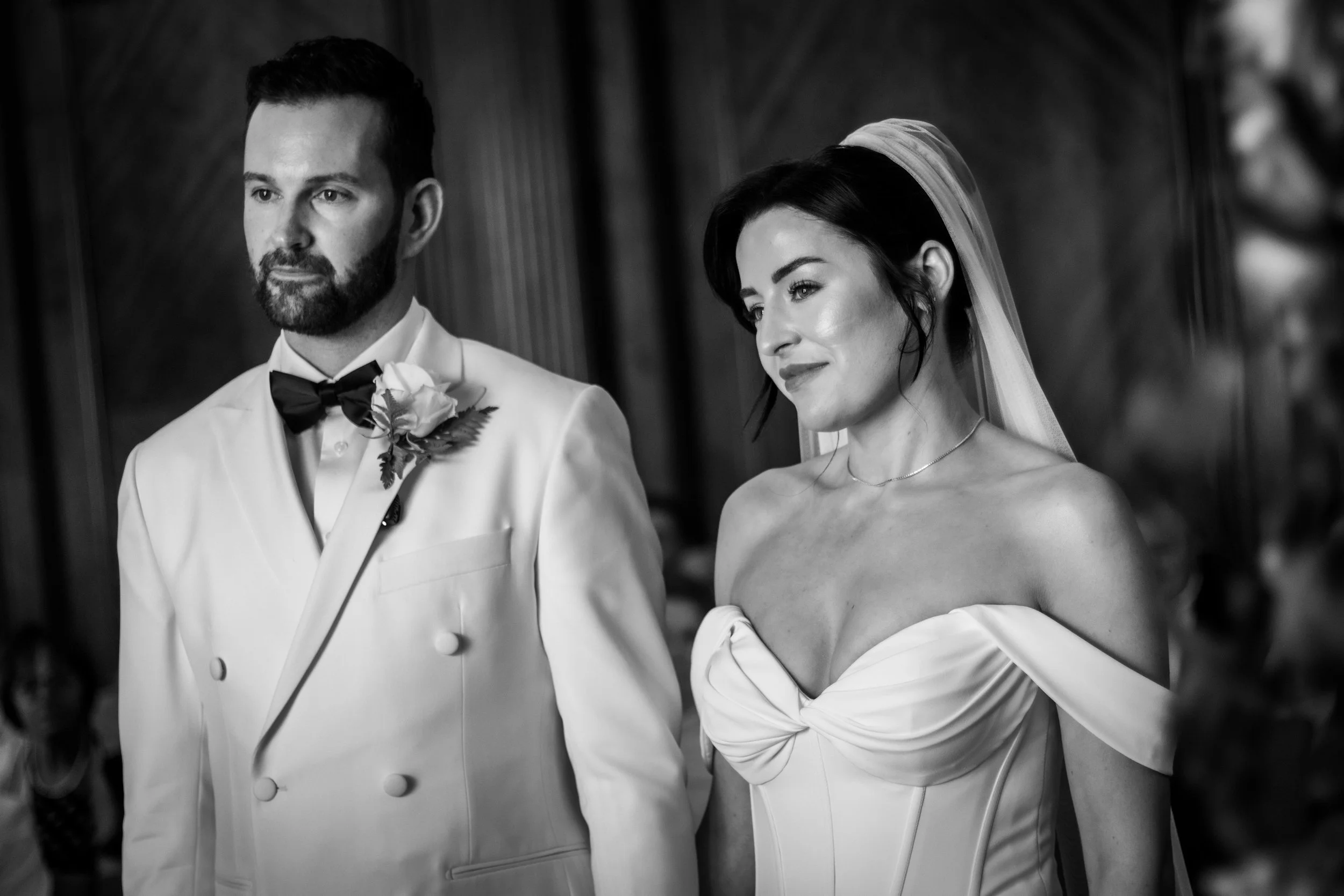 A black and white photo of a bride and groom standing together during a wedding ceremony. The groom is wearing a white tuxedo with a black bow tie and a boutonniere, while the bride is in an off-shoulder wedding gown with a veil.