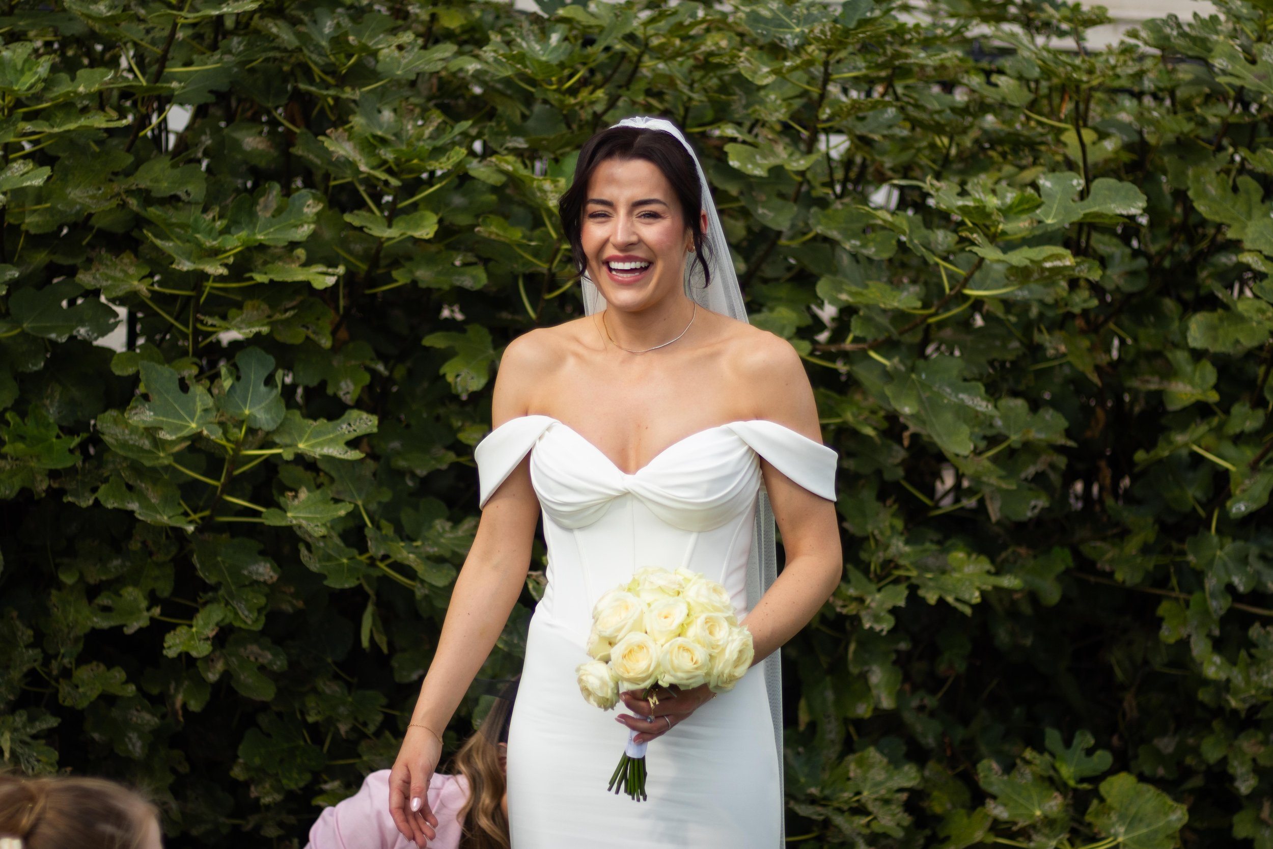 A smiling bride in a white wedding dress holding a bouquet of white roses, standing outdoors in front of green foliage.