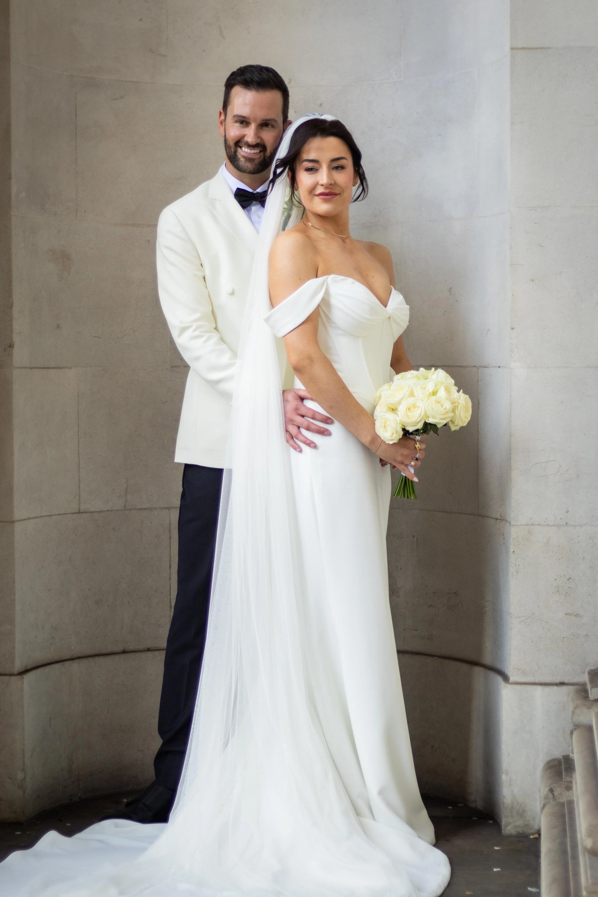 A bride and groom in wedding attire, standing close together with the groom's arm around the bride, both smiling. The bride holds a bouquet of white roses. They are indoors against a stone wall.