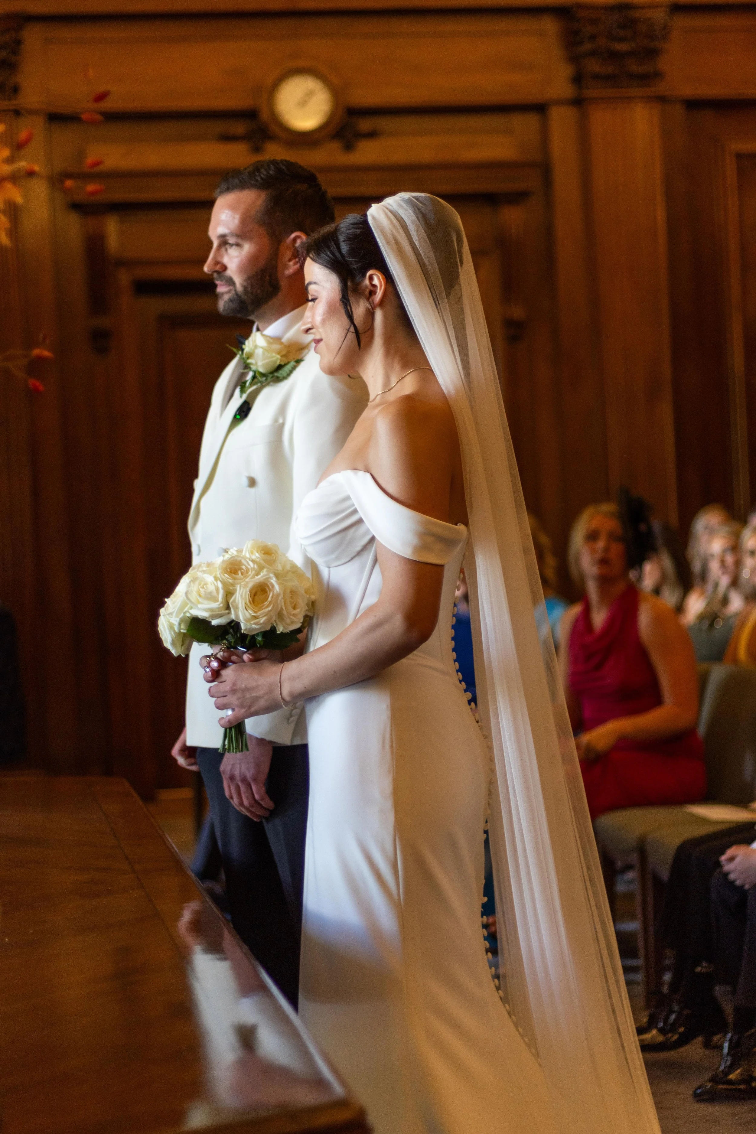A bride and groom standing side by side during their wedding ceremony, with the bride holding a bouquet of white roses and the groom dressed in a white tuxedo, in a room with wooden walls and guests seated nearby.