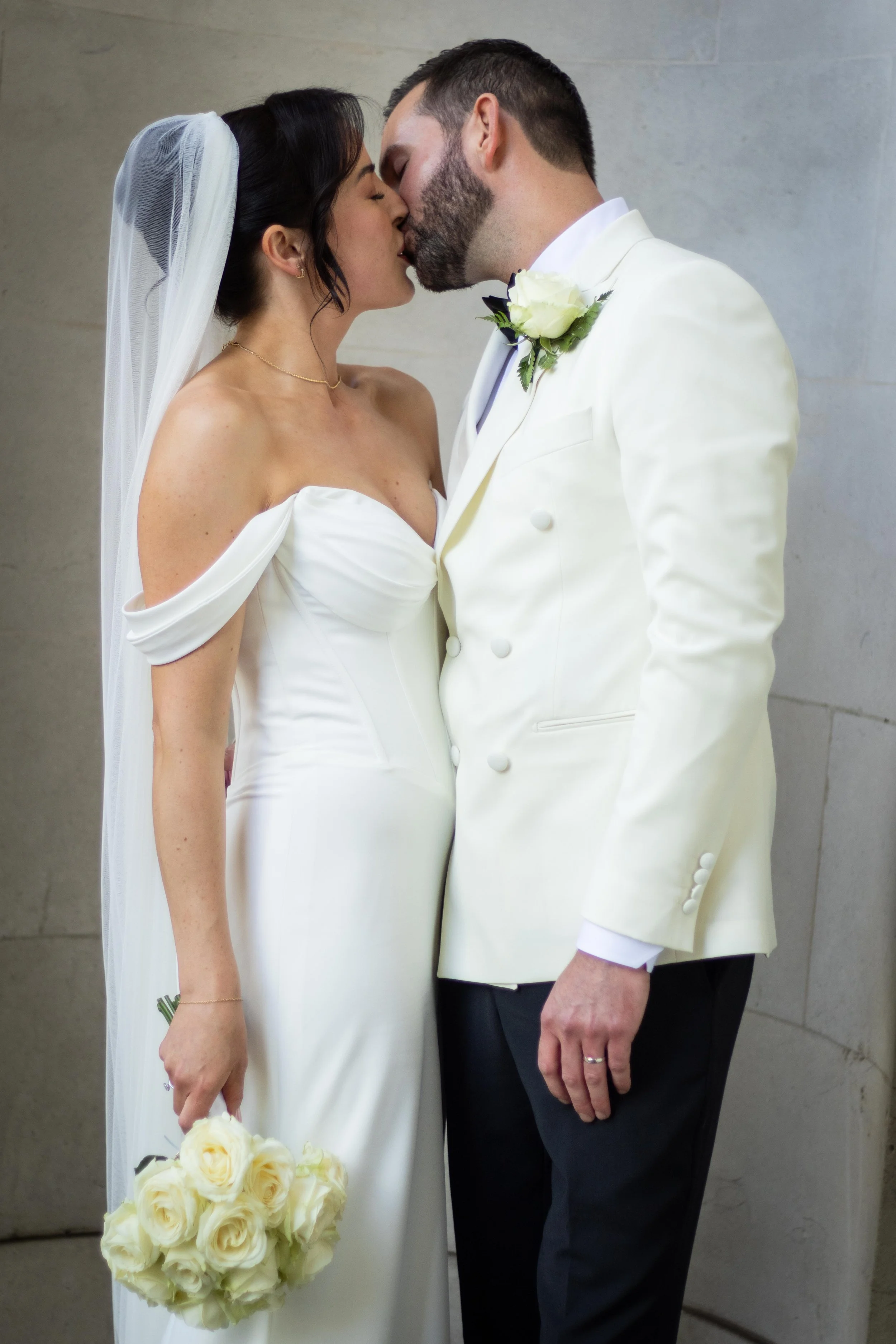 A bride and groom are kissing during their wedding, with the bride holding a bouquet of white roses.