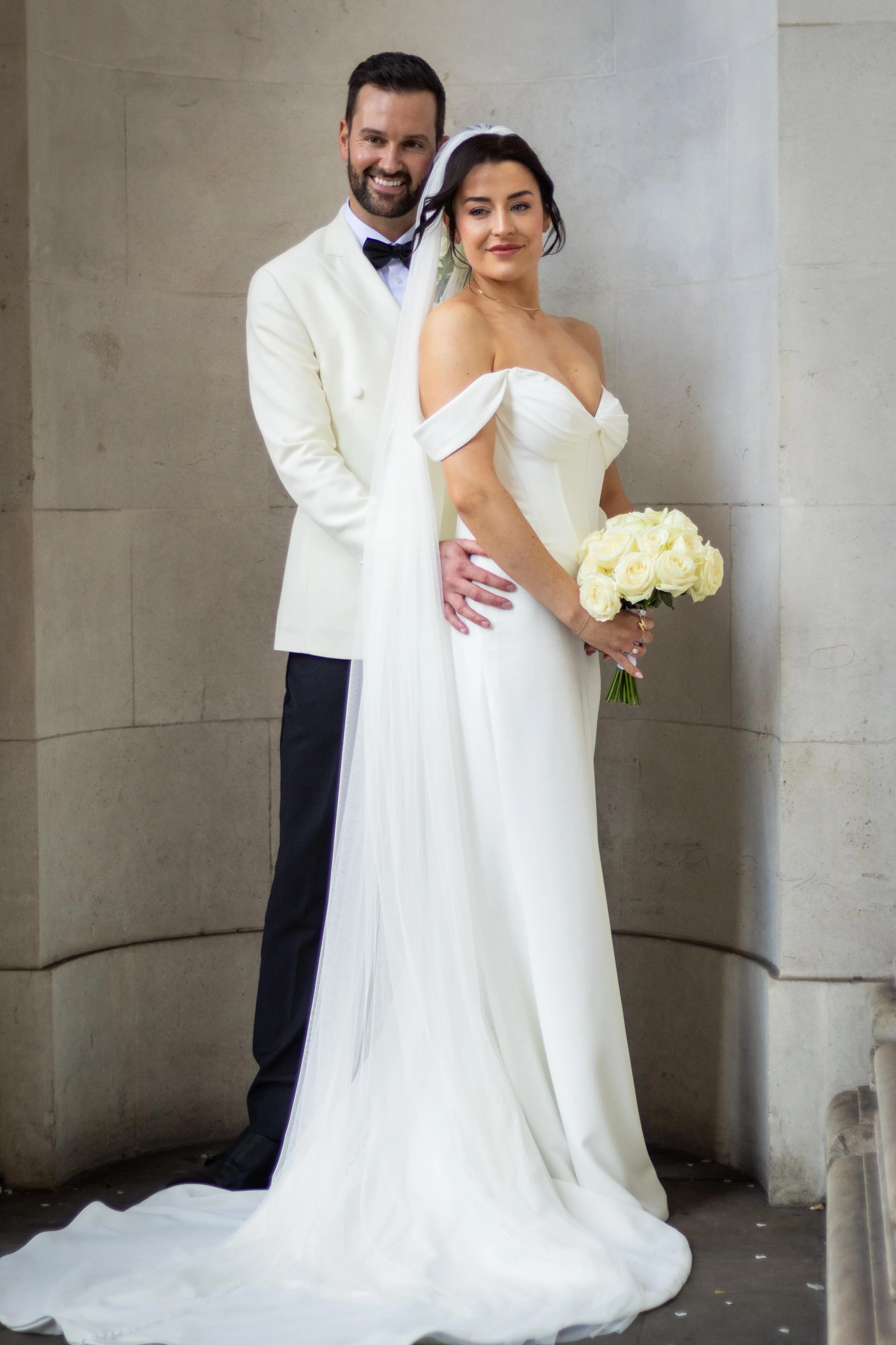 A newly married bride in an elegant white wedding dress holding a bouquet of white roses, standing in front of a neutral-colored wall, with a smiling groom in a white tuxedo with a black bow tie standing behind her.