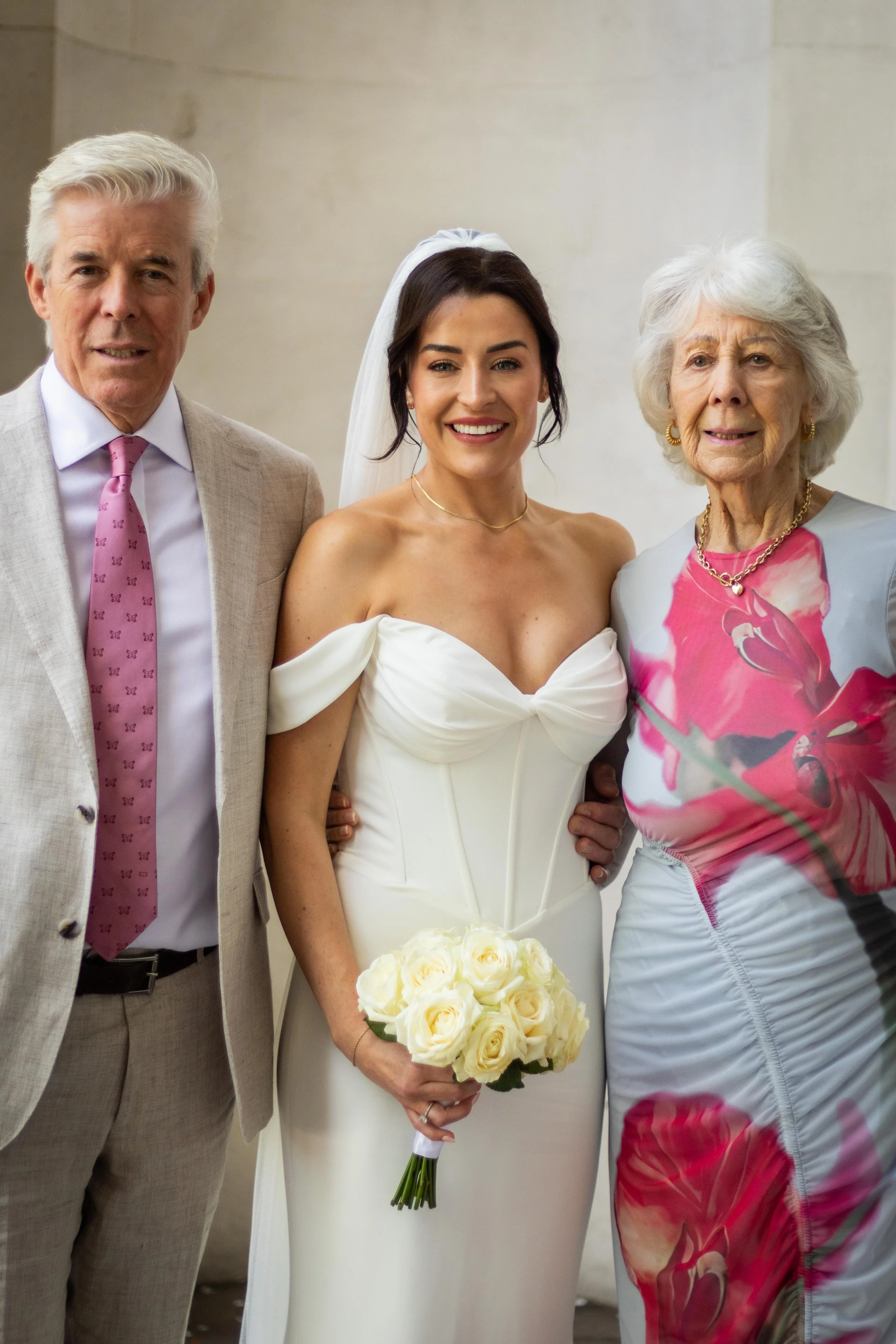 A bride in a white wedding dress holding a bouquet of white roses stands between an older man in a beige suit with a pink tie and an older woman in a colorful dress, all smiling.