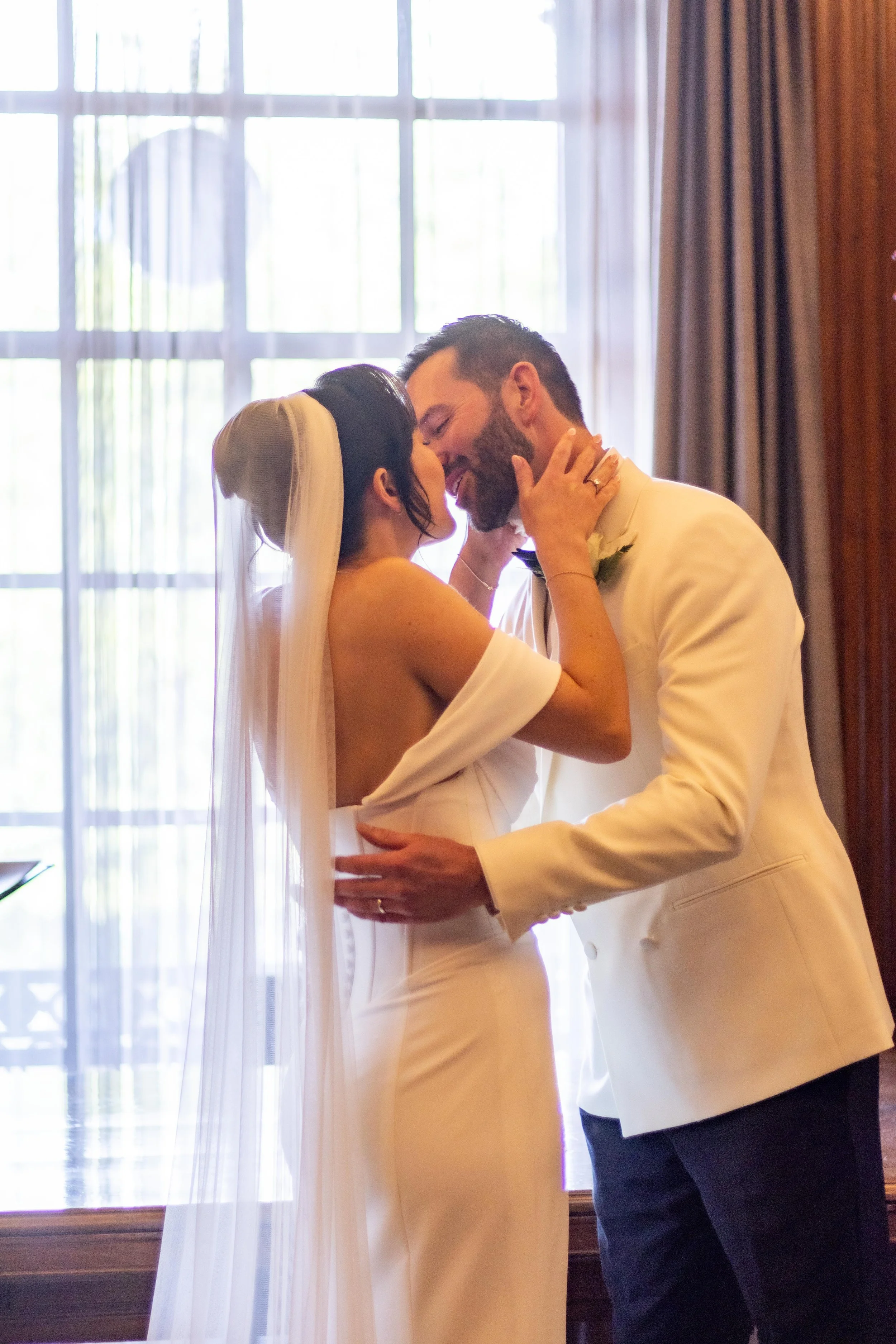 A bride and groom sharing a kiss indoors, with the bride wearing a white off-shoulder wedding dress with a veil, and the groom in a white tuxedo jacket, against a large window with curtains.