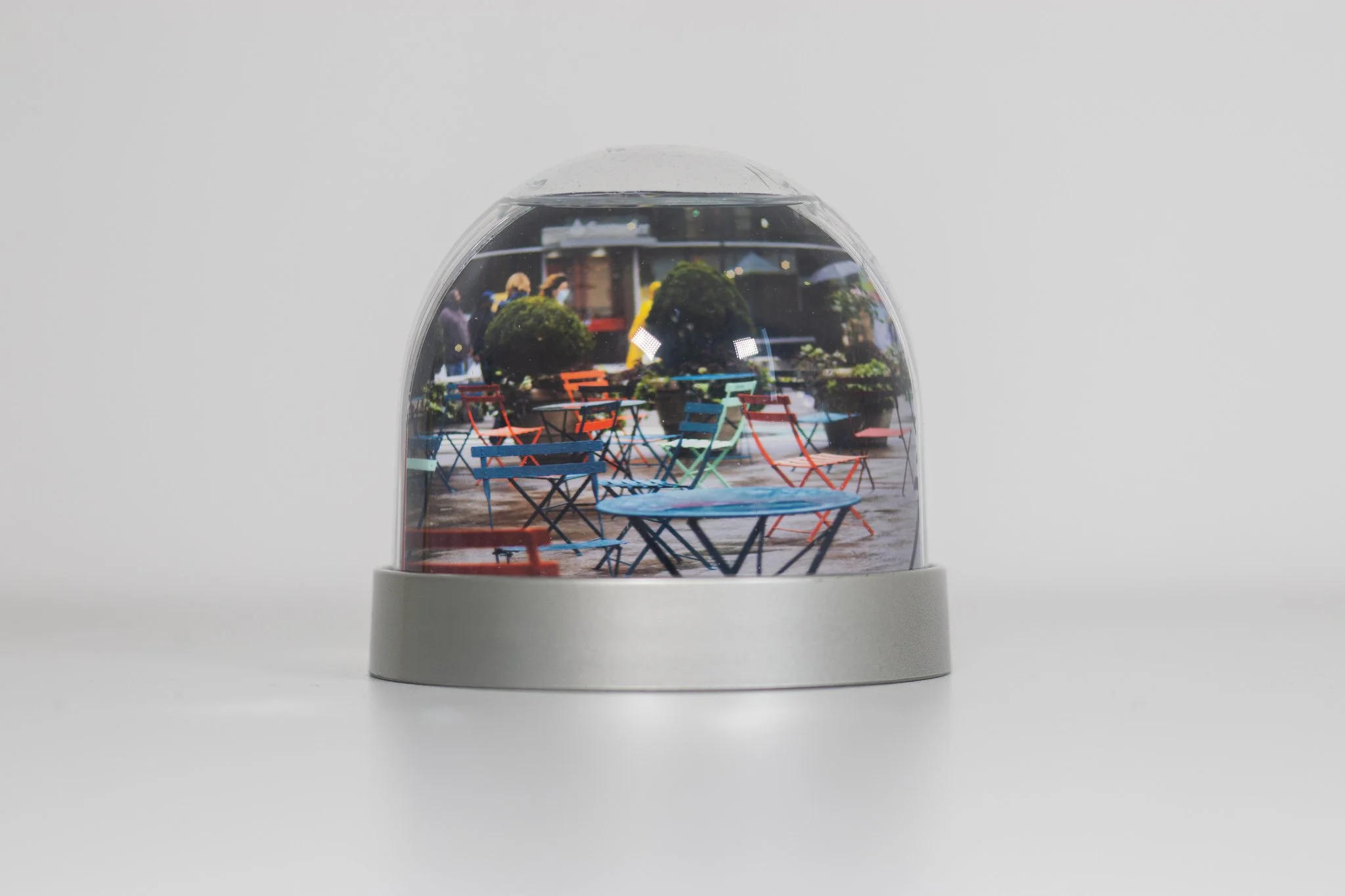 A snow globe with a miniature outdoor café scene featuring colorful tables and chairs, with a city street background reflected in the glass.