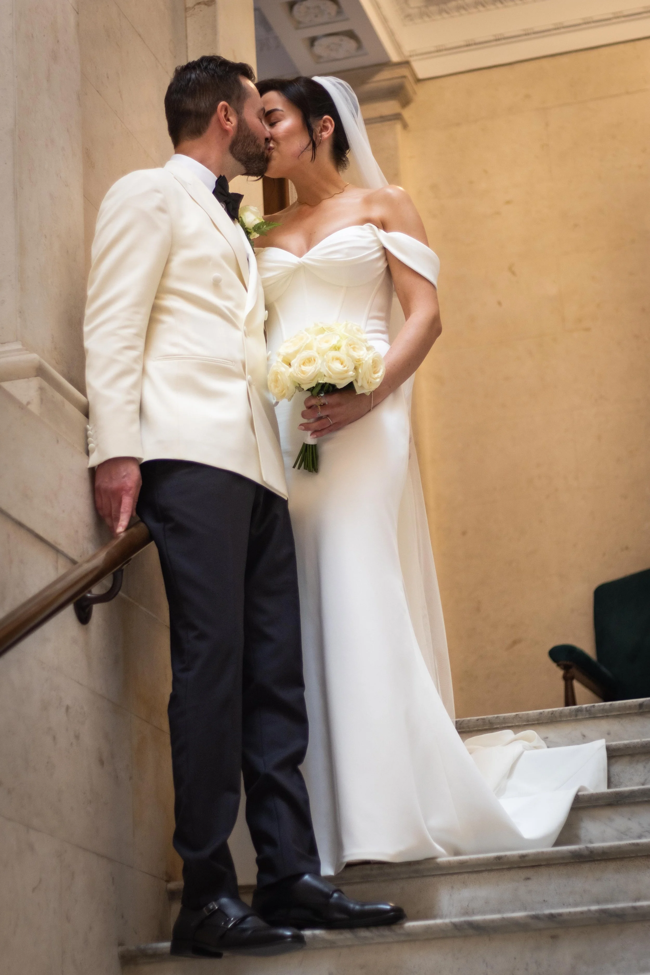 A bride and groom sharing a kiss on the staircase of a formal venue. The bride is in a white off-shoulder wedding dress holding a bouquet of white roses, and the groom is in a cream tuxedo jacket with black trousers.