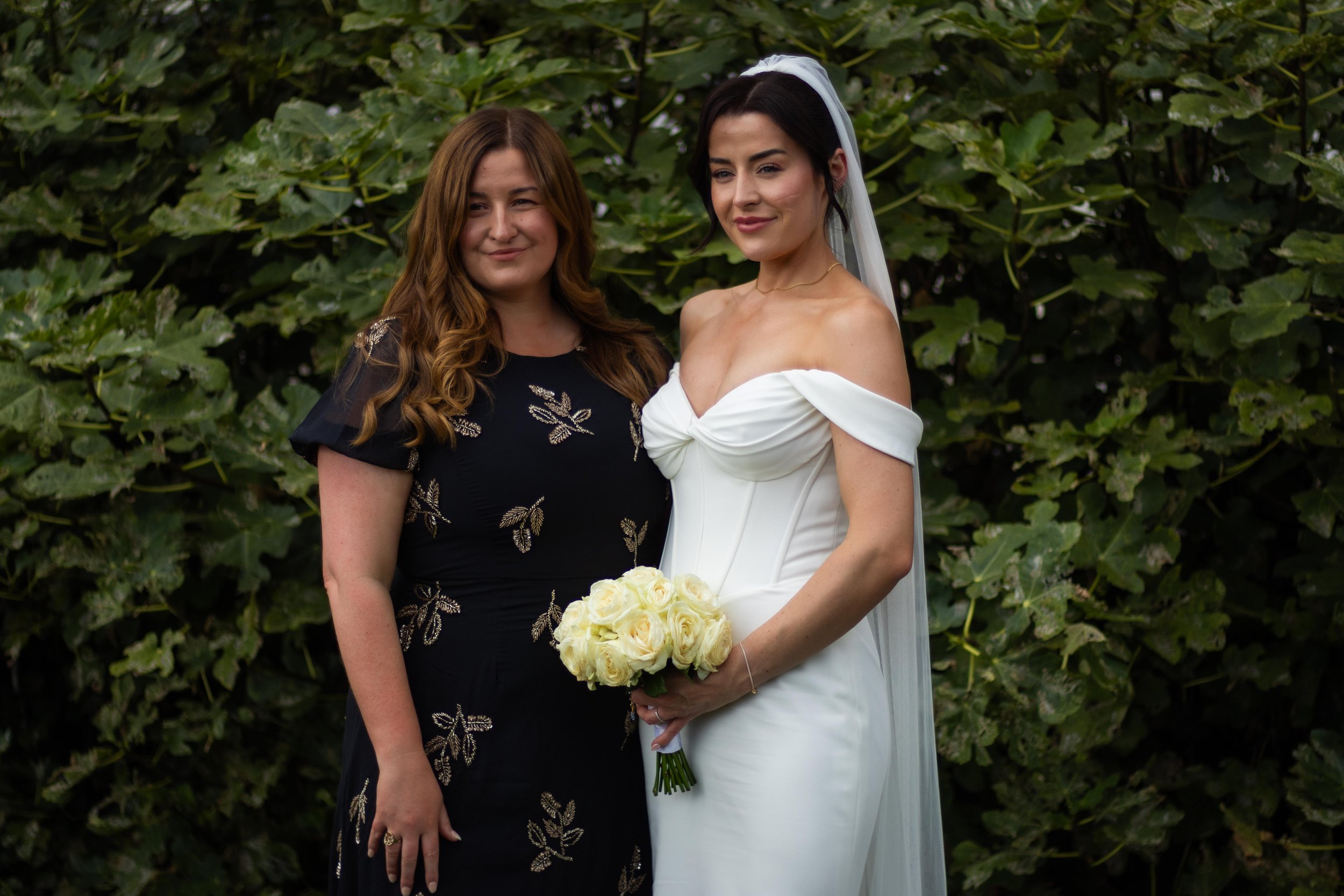 A bride in a white off-shoulder wedding gown and veil holding a bouquet of white roses, standing beside a woman in a black dress with gold embroidery, in front of green foliage.