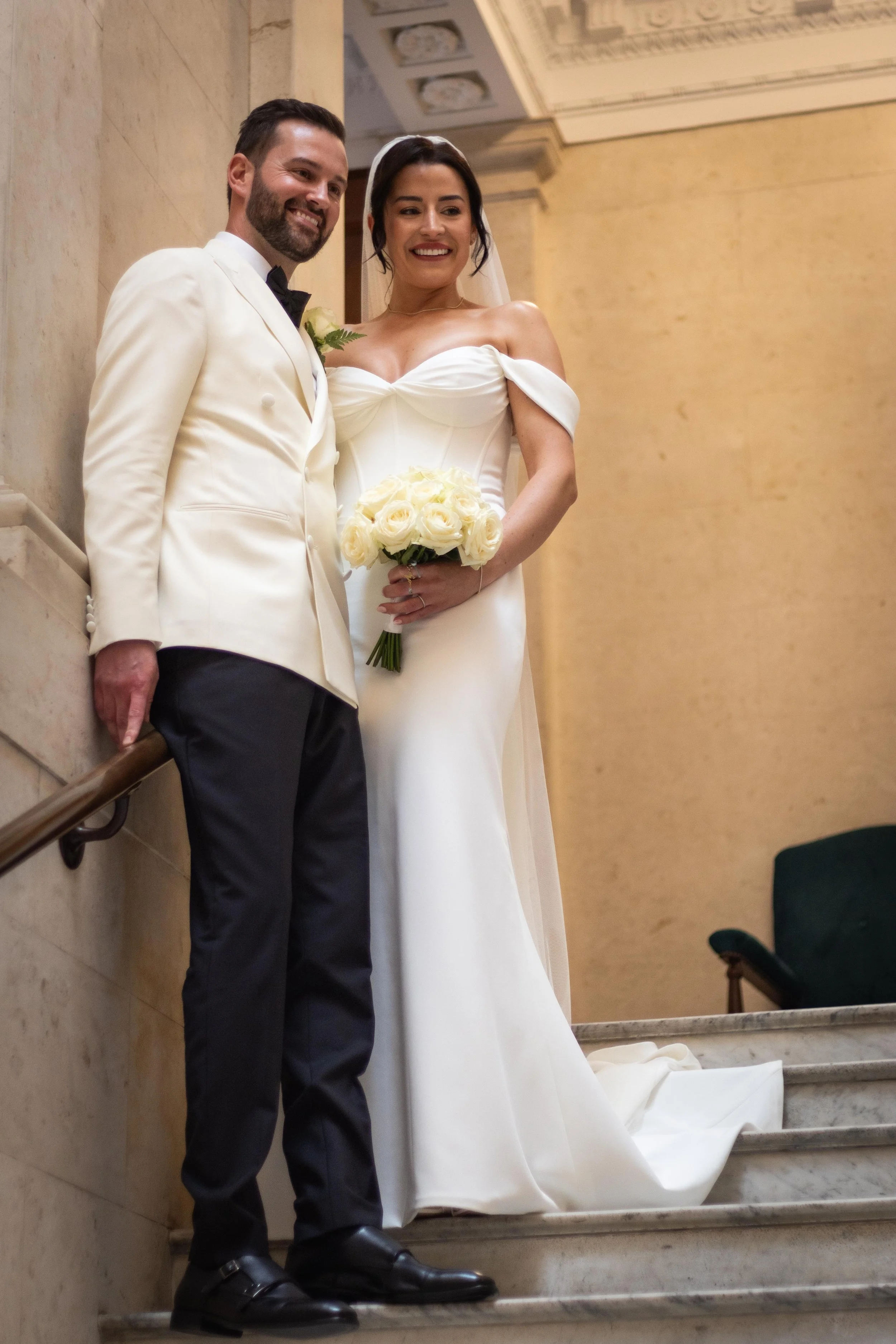A newlywed couple standing on a staircase inside a building, smiling, with the bride holding a bouquet of white roses.